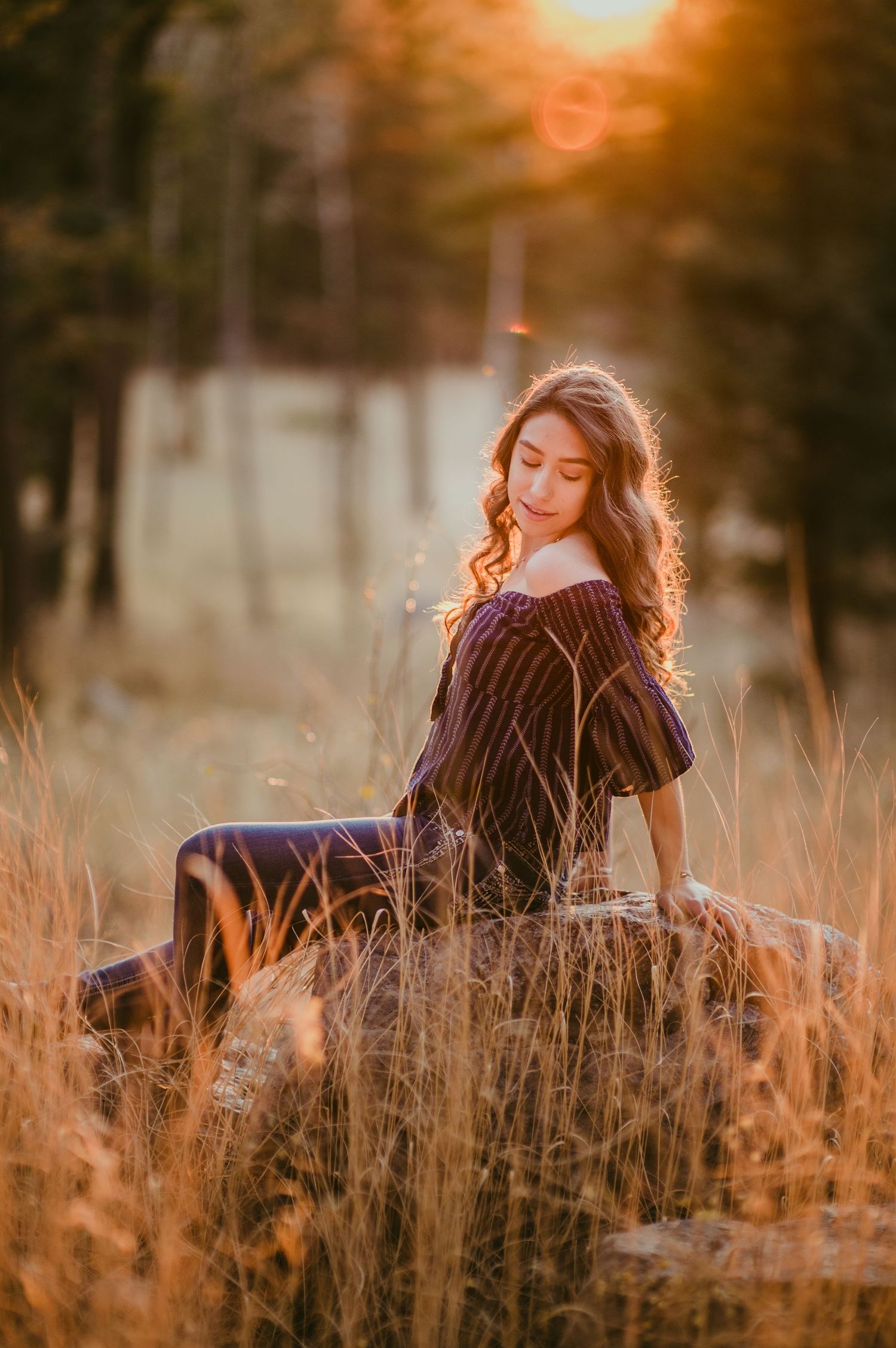 Young woman in off-shoulder dress sitting on rock in golden sunset light surrounded by tall grass and trees.