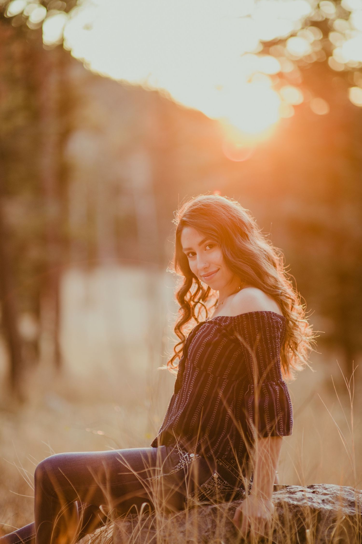 Young woman with long wavy hair sits outdoors in golden sunset light, wearing an off-shoulder dark dress in a field.