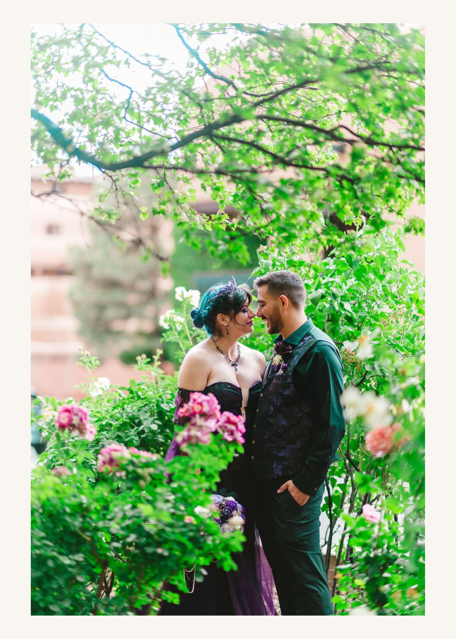 A couple in elegant wedding attire shares a romantic moment surrounded by lush green foliage and pink blooming flowers.