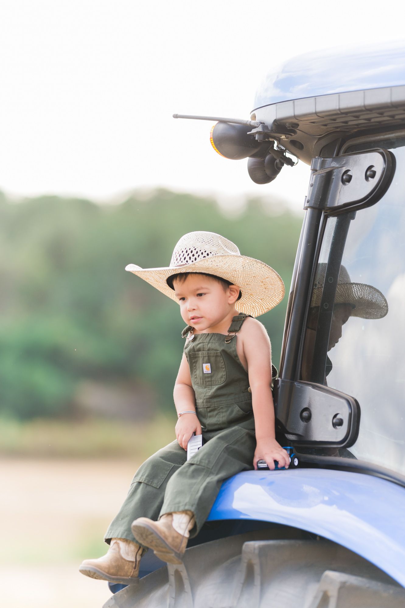 Toddler in overalls and straw cowboy hat sitting on a blue tractor in a farm field.