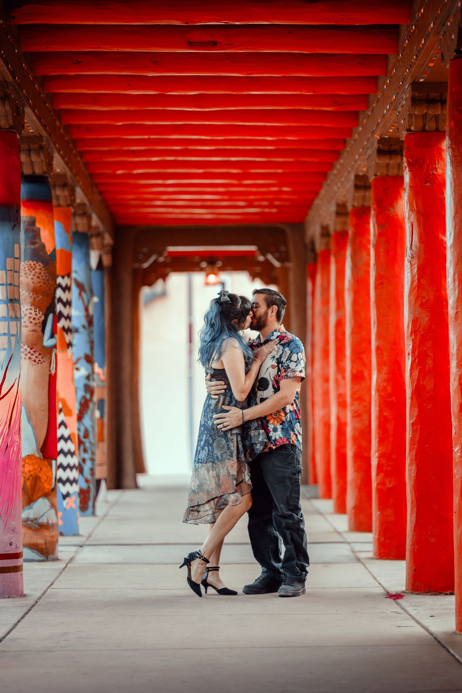 Couple sharing a romantic kiss under vibrant red columns of an ornate corridor with colorful artwork.