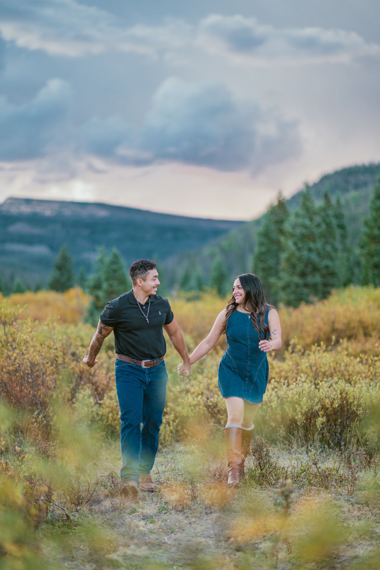Couple holding hands walking through colorful autumn meadow with mountain backdrop and dramatic cloudy sky.