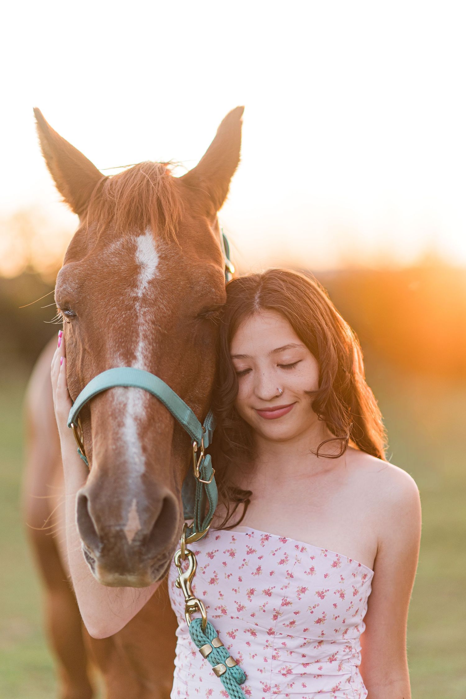 A chestnut horse with a white blaze stands close to someone wearing a floral dress in warm sunset lighting.