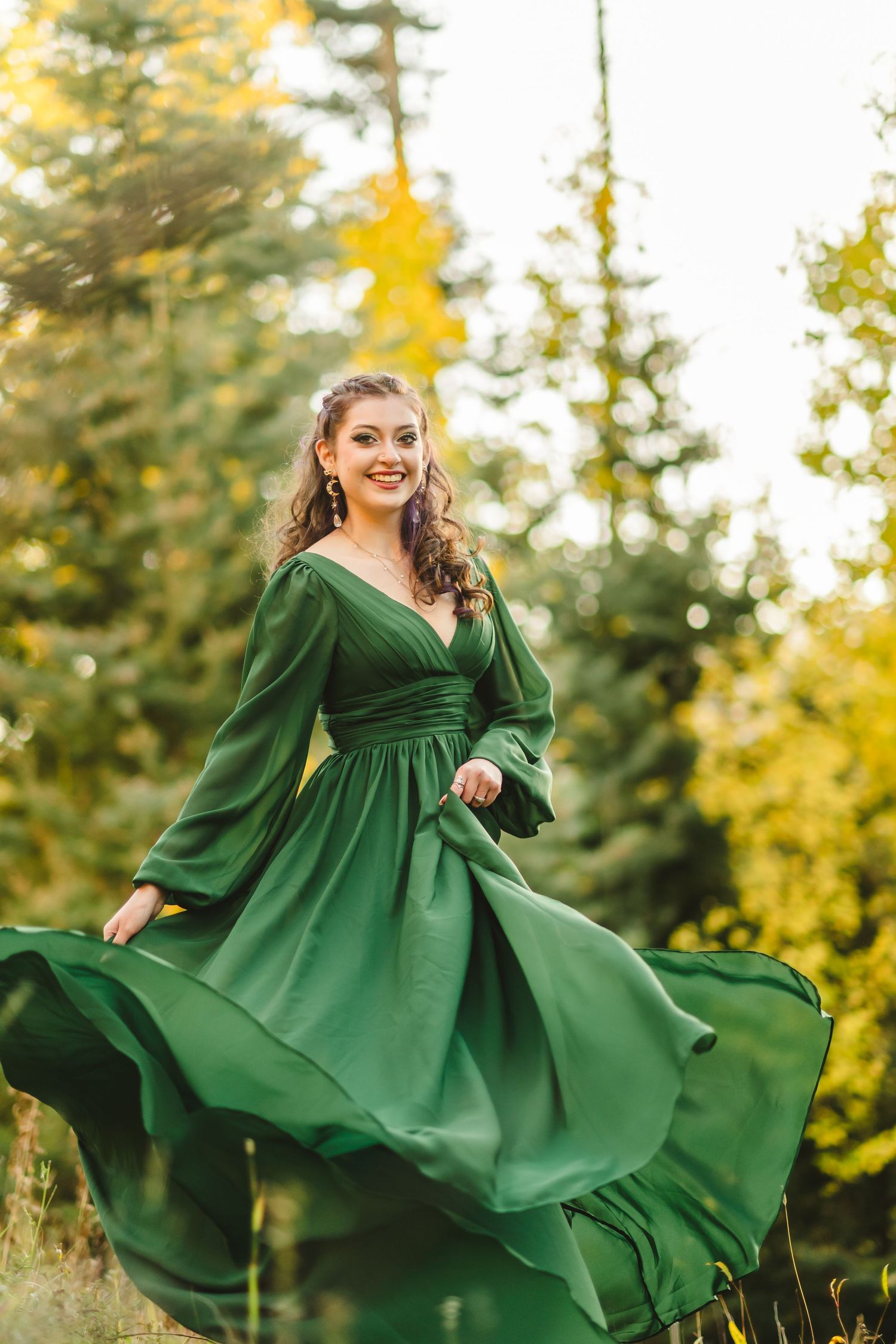 A flowing emerald green dress twirls gracefully in an outdoor autumn setting with golden foliage in the background.