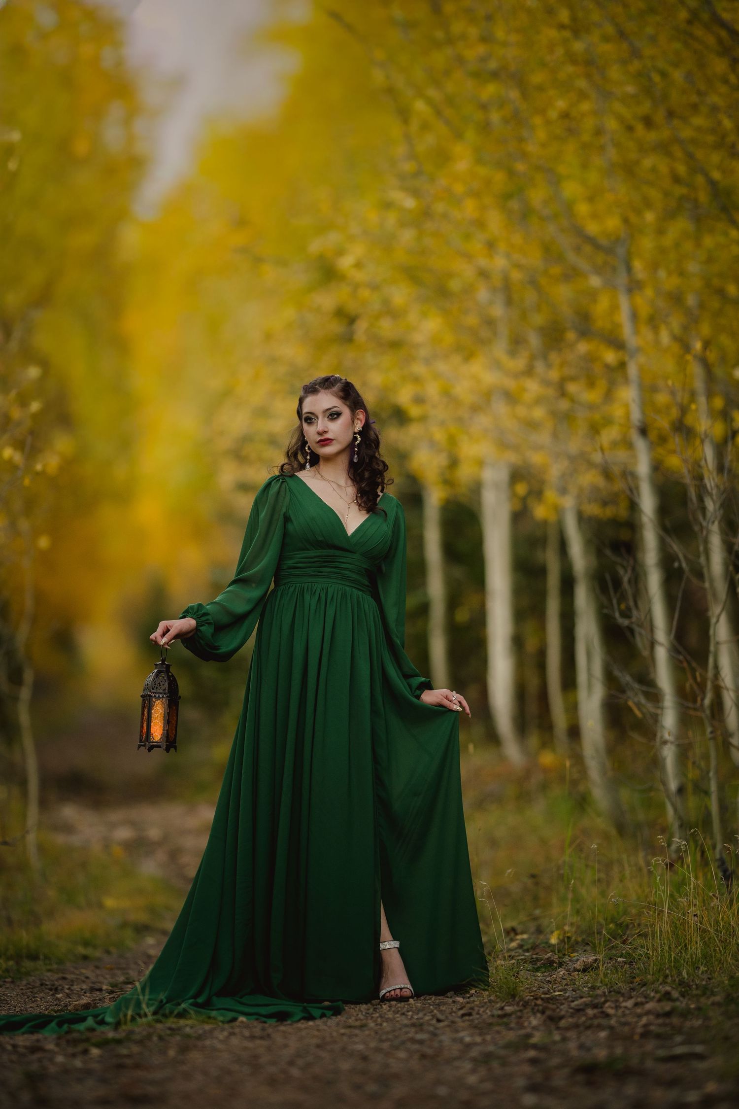 Elegant emerald green dress flows in an autumn forest path with golden aspen trees and a vintage lantern.