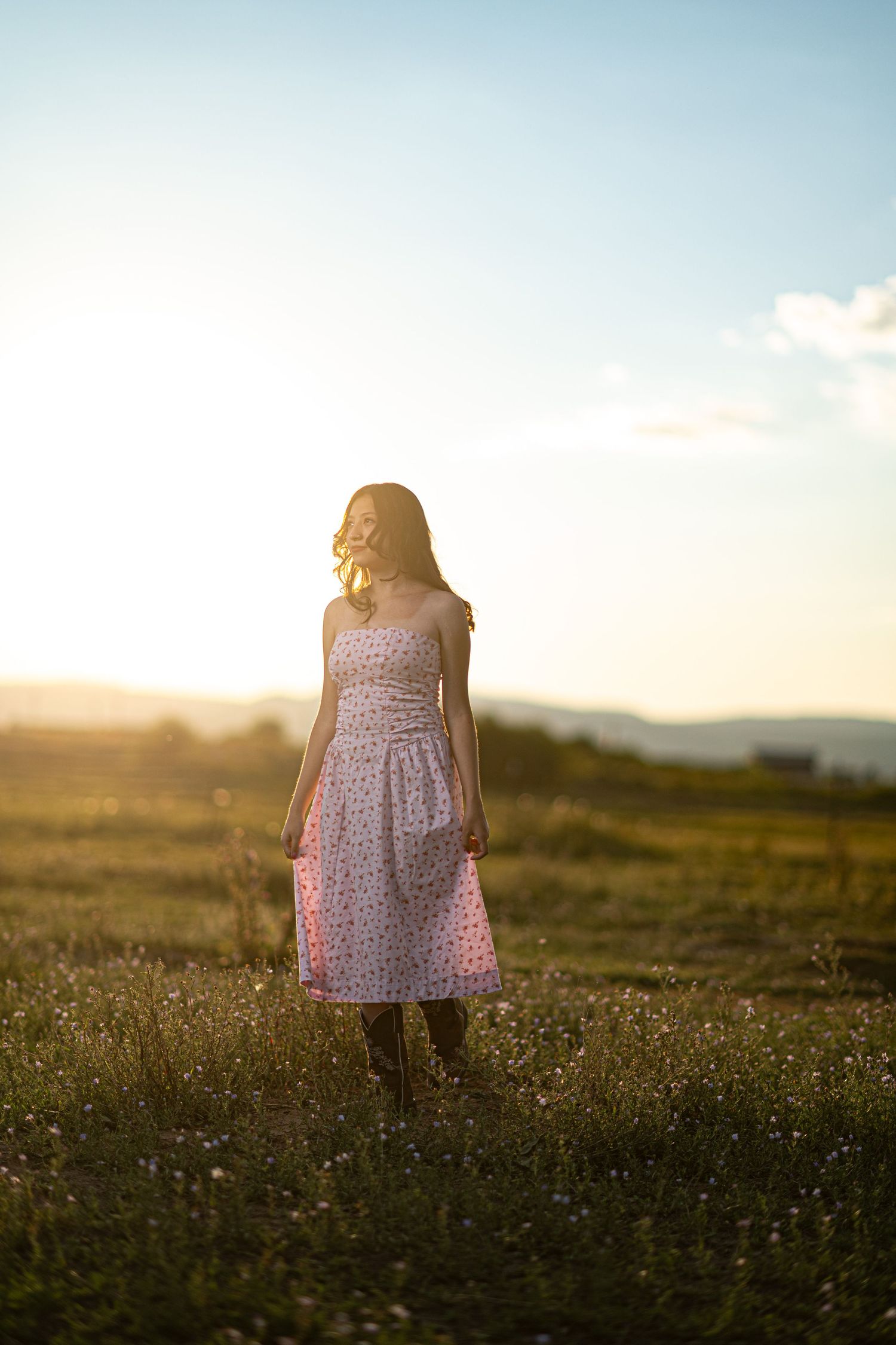 Someone wearing a floral sundress stands in a golden meadow at sunset with soft light illuminating the wildflowers.