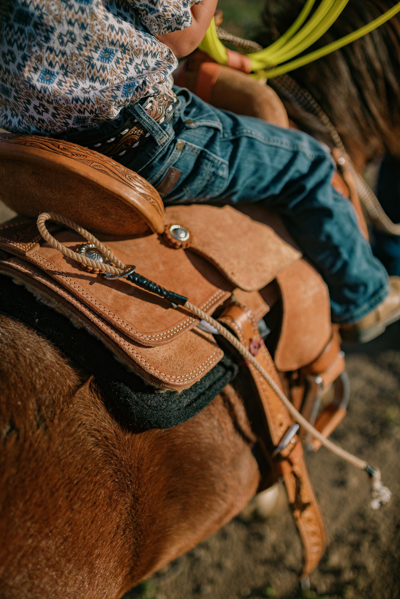Close-up of a Western leather saddle on a brown horse with a rider in jeans holding a lasso rope.