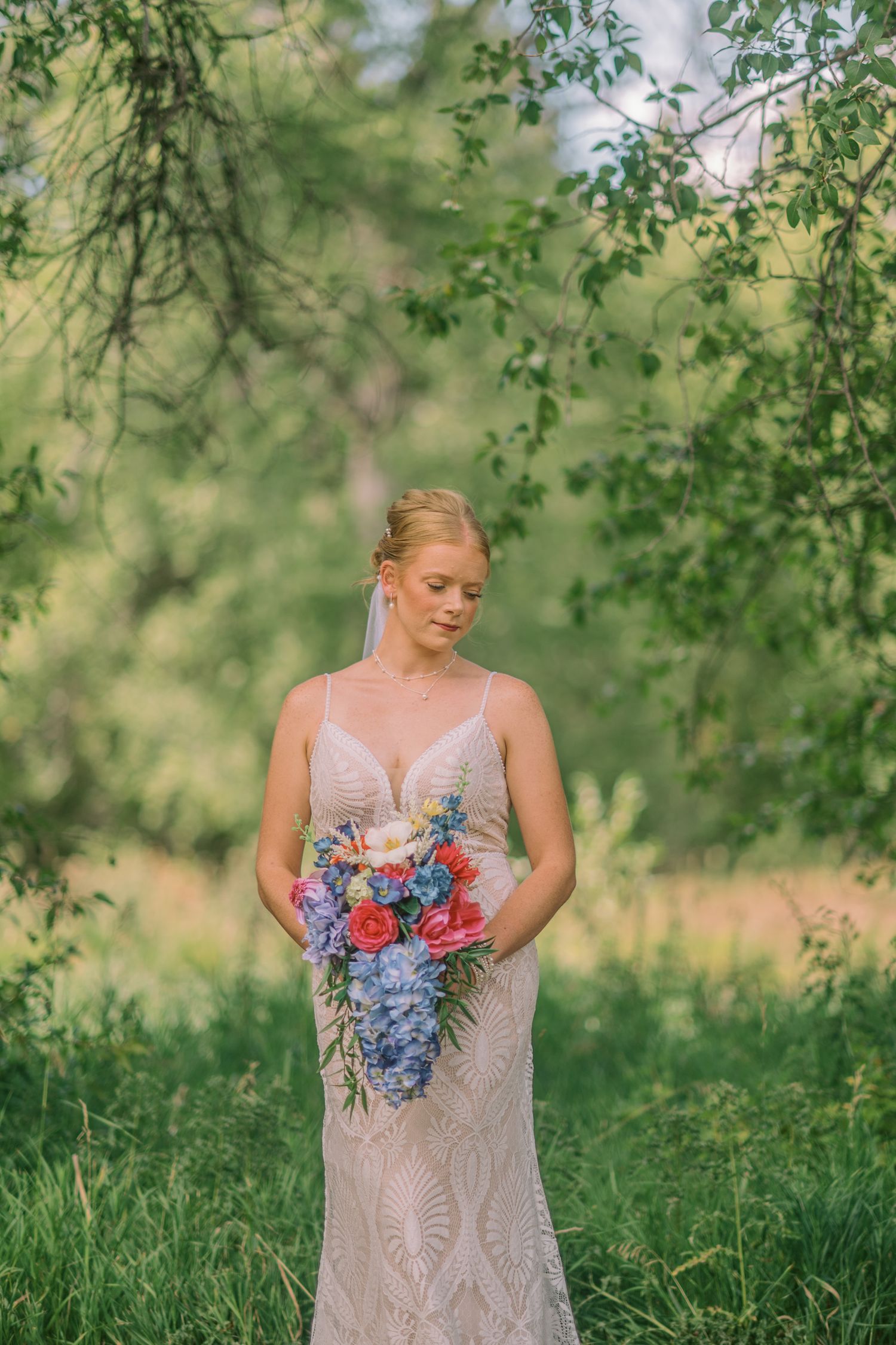 Bride in lace gown holds colorful bouquet of red and blue flowers outdoors under green trees.
