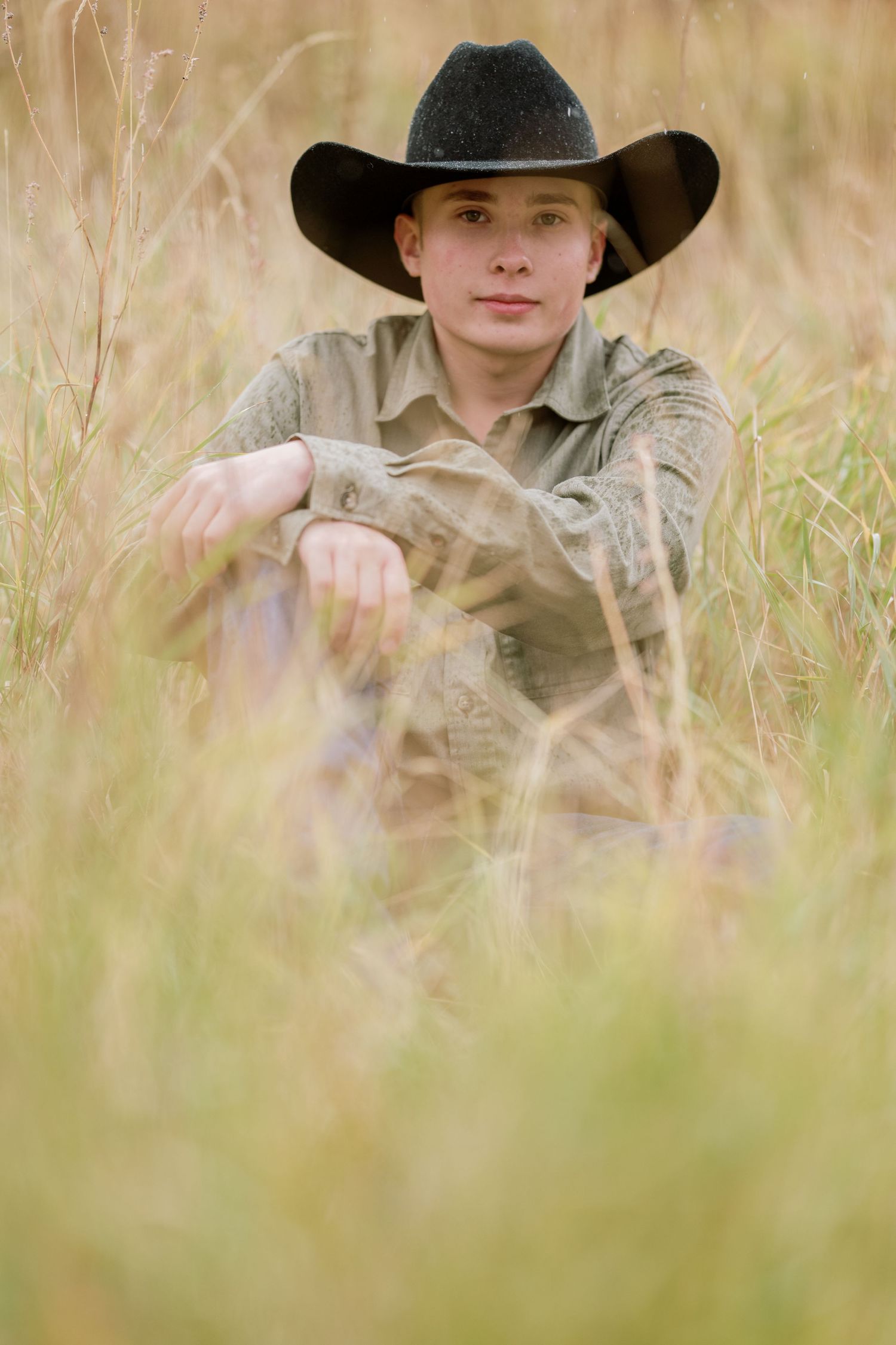 A person wearing a black cowboy hat and light colored shirt sits among tall prairie grass in a rustic outdoor setting.