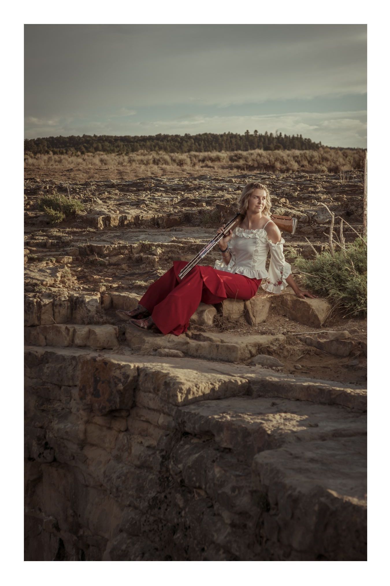 Woman in red skirt playing violin on ancient ruins in a desert landscape under cloudy skies.