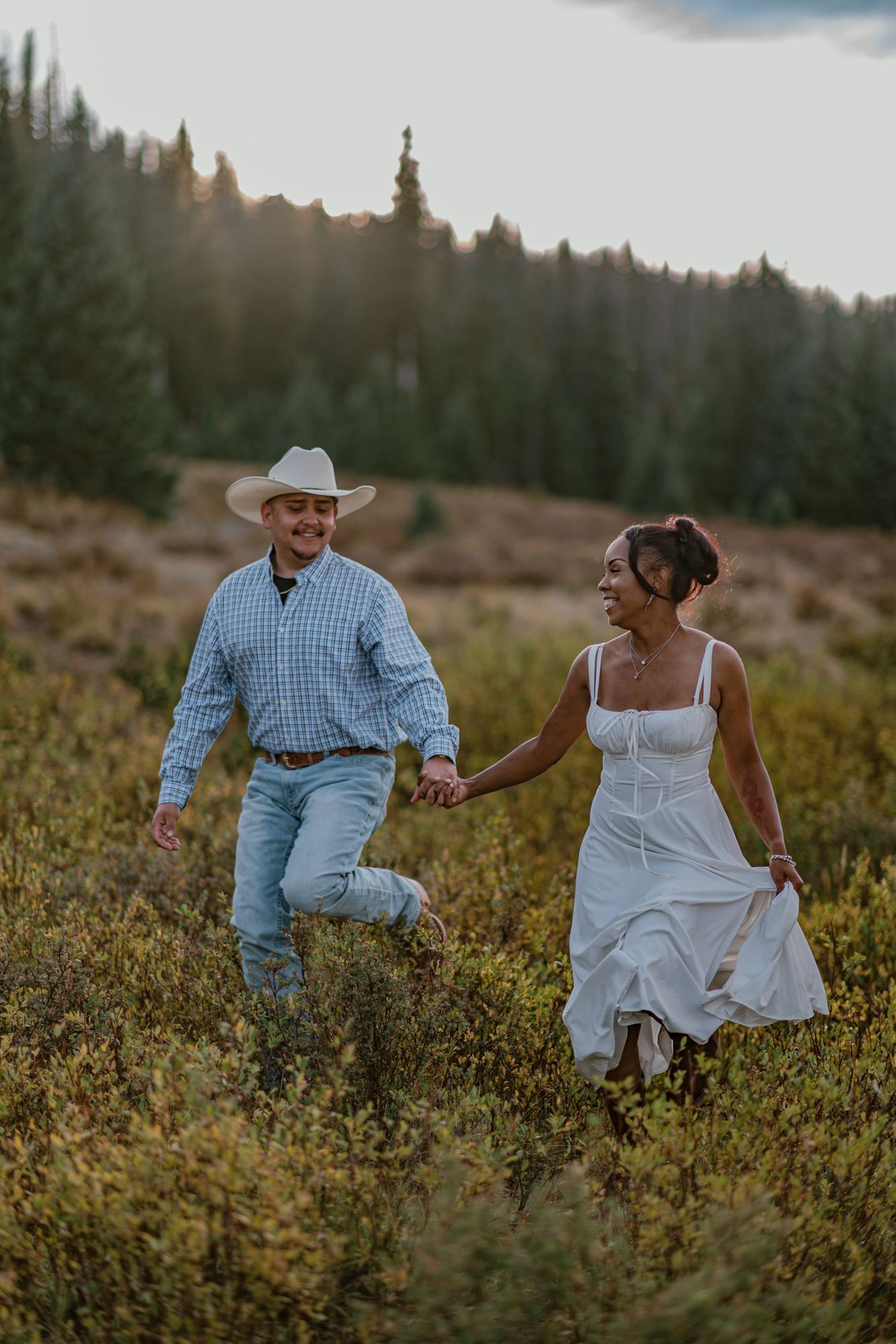 A joyful couple runs hand-in-hand through a wildflower meadow at sunset, man in cowboy hat and woman in white dress.