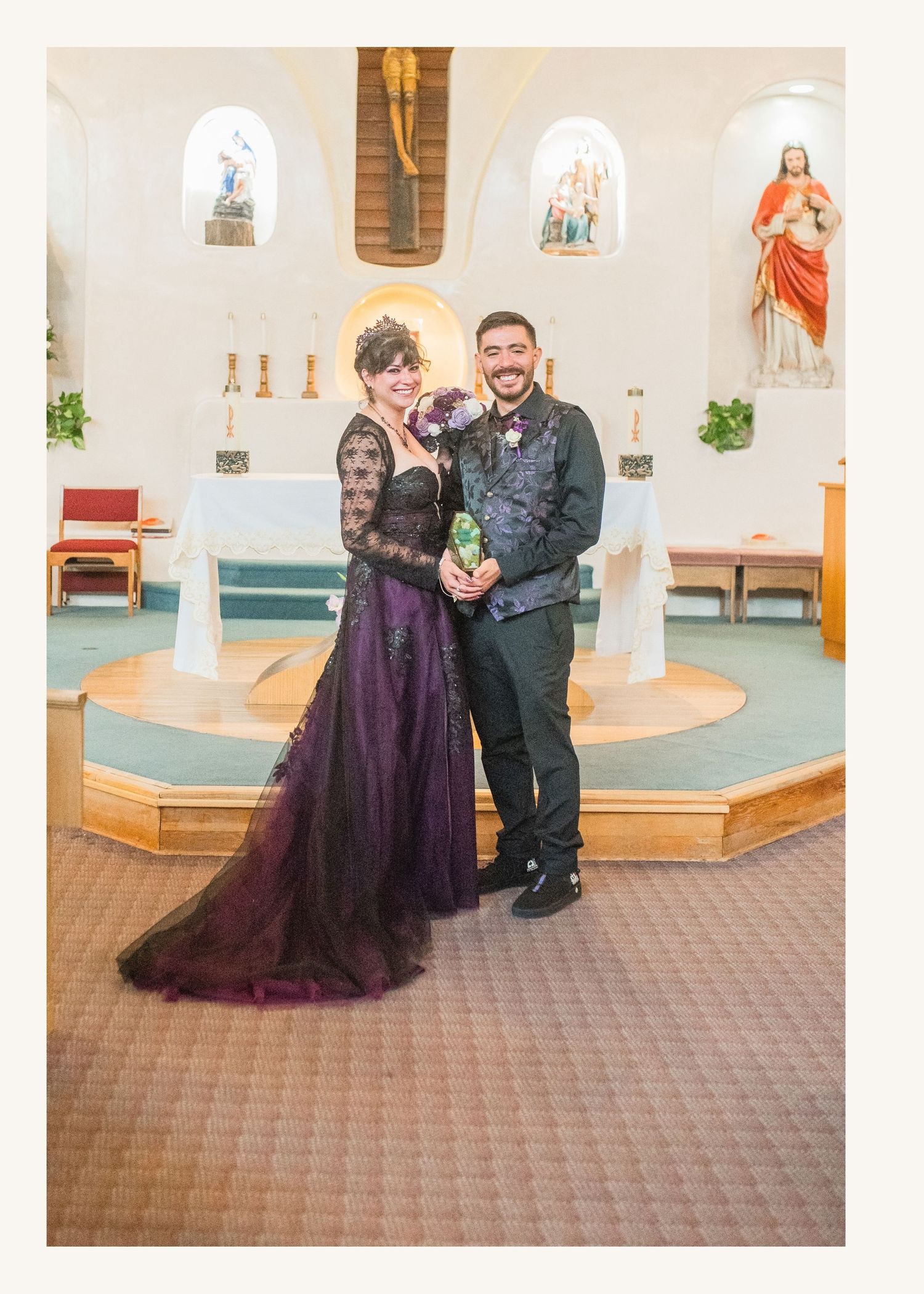 Couple in gothic wedding attire poses in church, bride in purple gown, groom in black suit, altar backdrop.