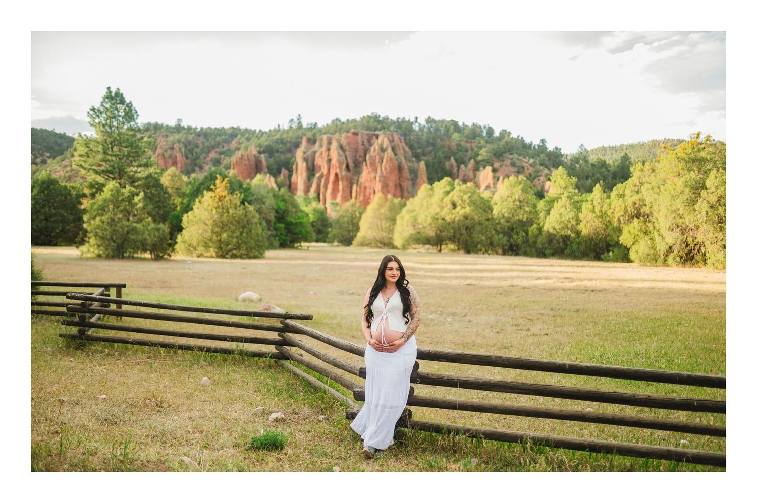 Pregnant woman in white dress posing by wooden fence in scenic field with red rock formations and forest backdrop.