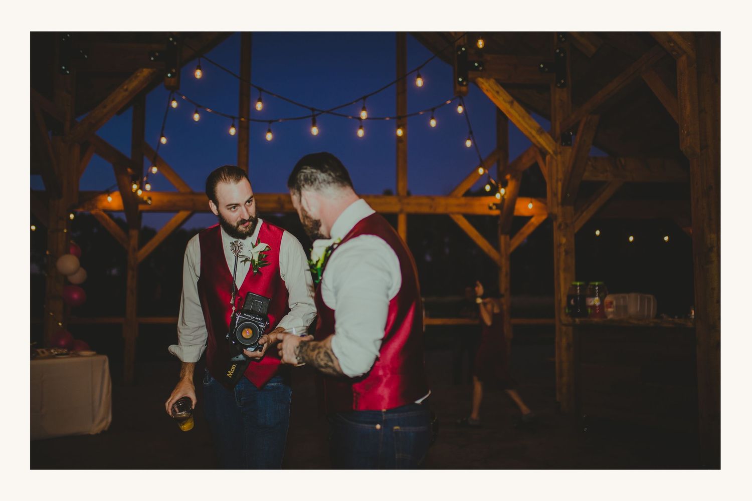 Two grooms in red vests examine a camera under string lights at an outdoor evening wedding reception.
