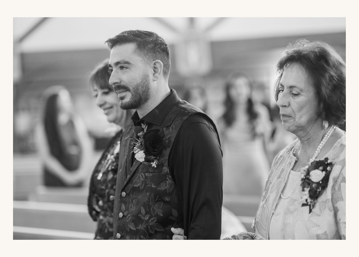 Black and white wedding photo of groom in floral vest standing with two women wearing corsages at ceremony.