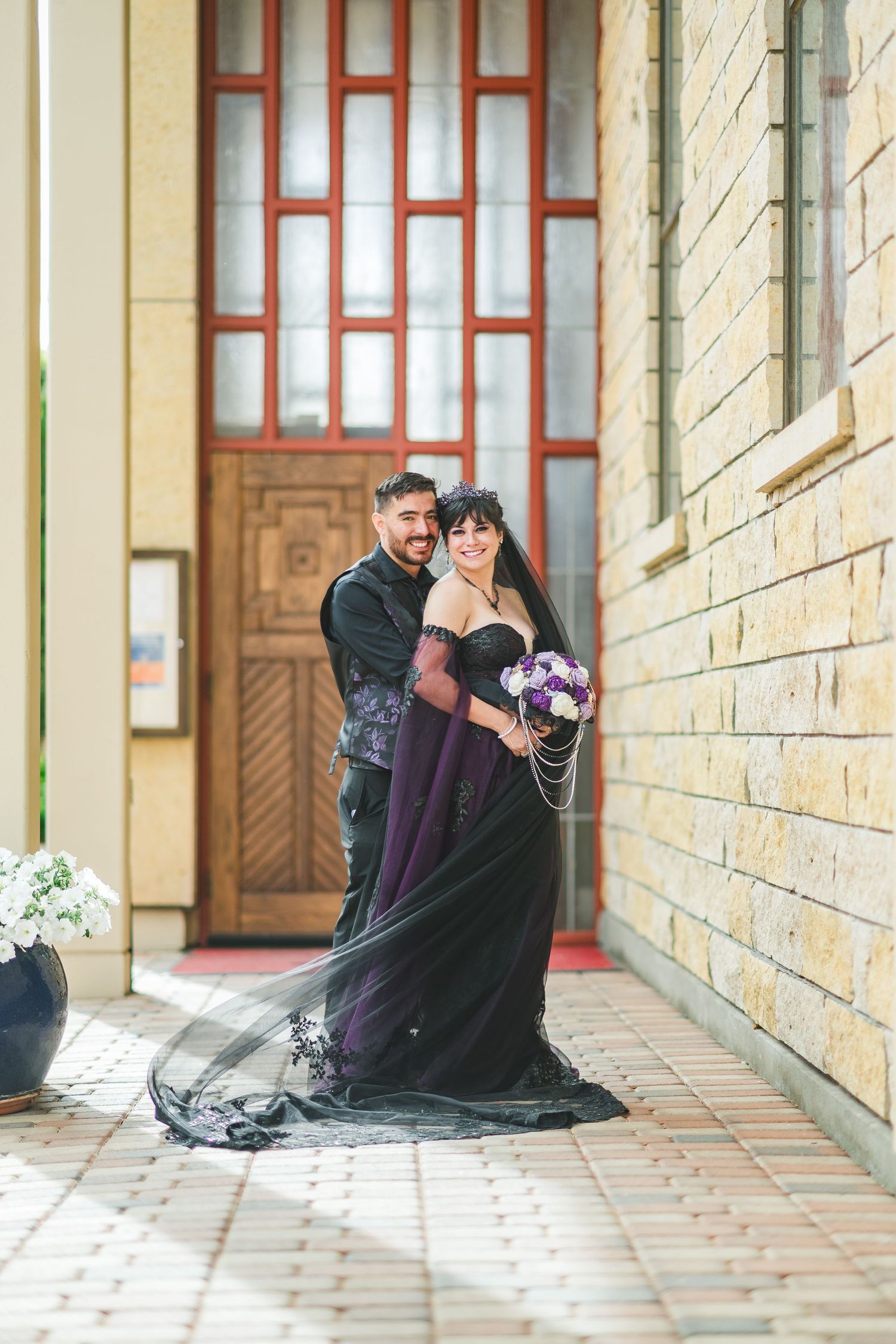 Couple in gothic wedding attire, bride in purple black gown with veil, groom in dark suit, posing before ornate wooden doors.