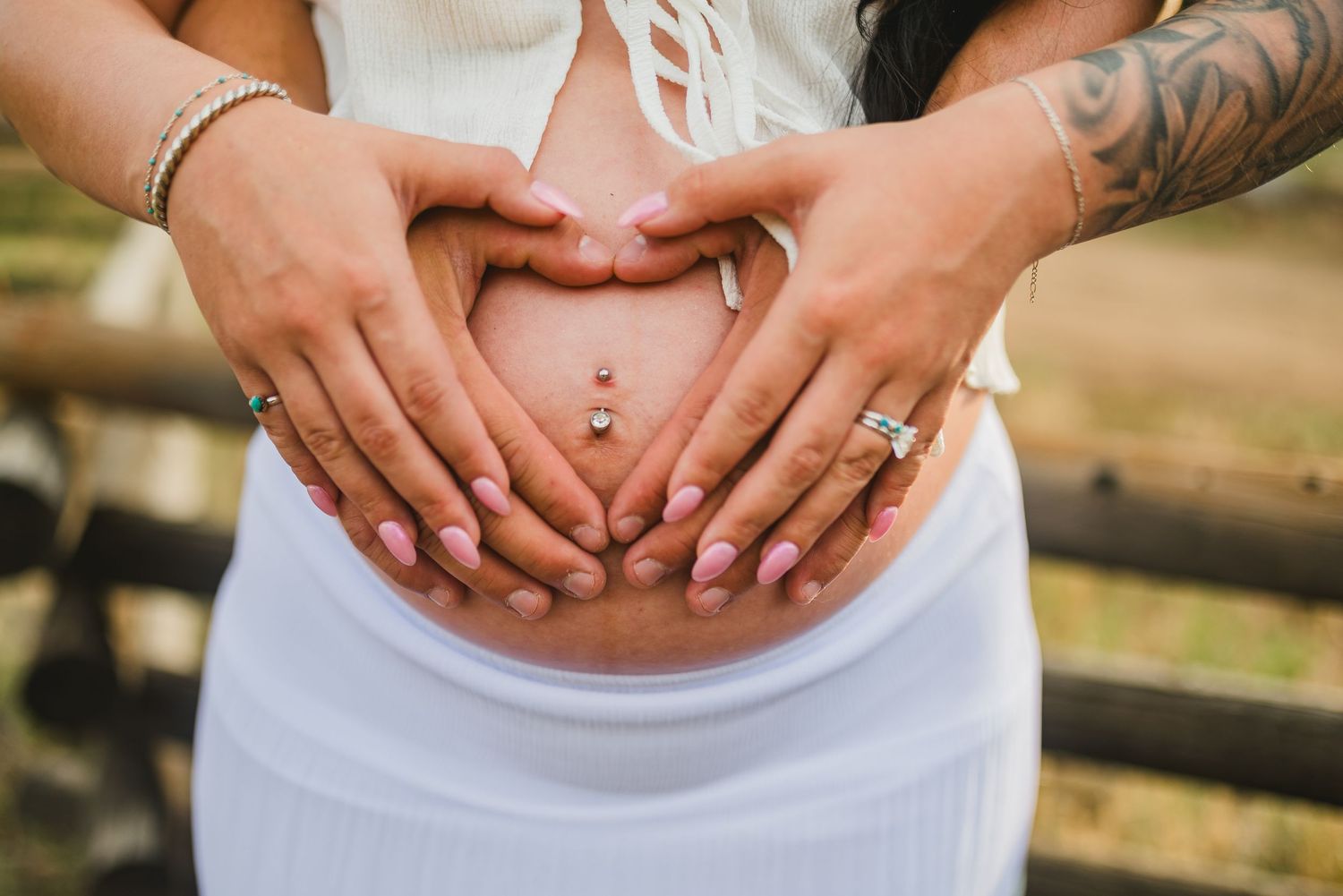 Couple forming heart shape with hands on pregnant belly, woman with pink nails and belly piercing wearing white skirt.