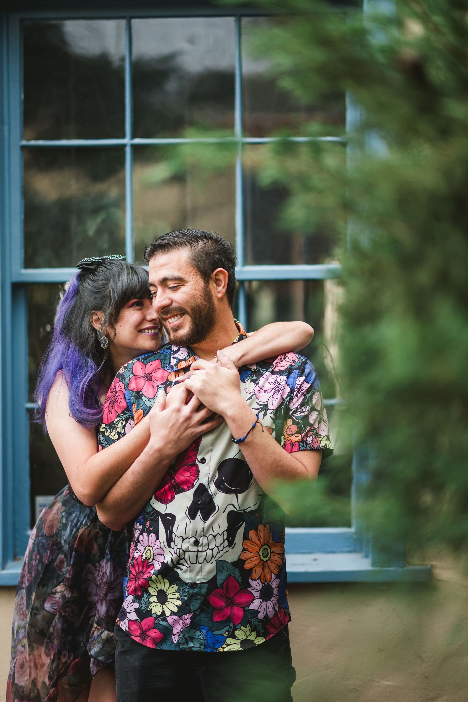 A couple embraces and smiles in front of a blue window, the man wearing a floral skull shirt and woman with purple hair.