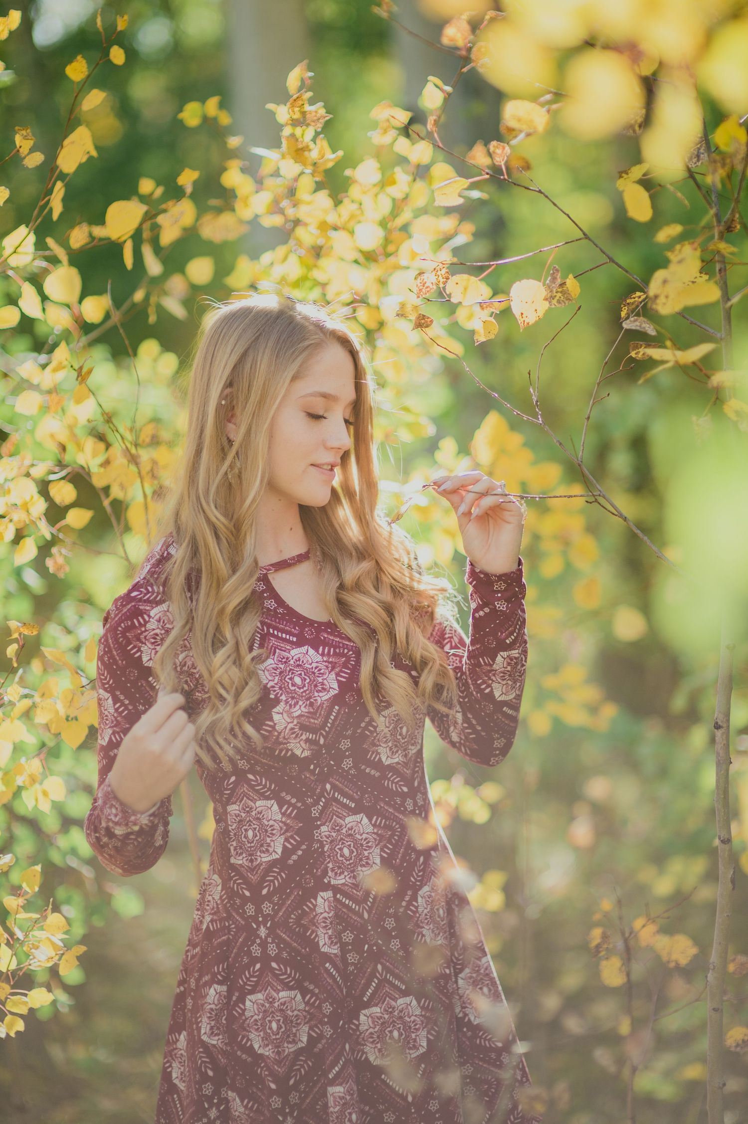 A romantic autumn scene with flowing blonde hair and a burgundy floral dress amongst golden yellow leaves and branches.