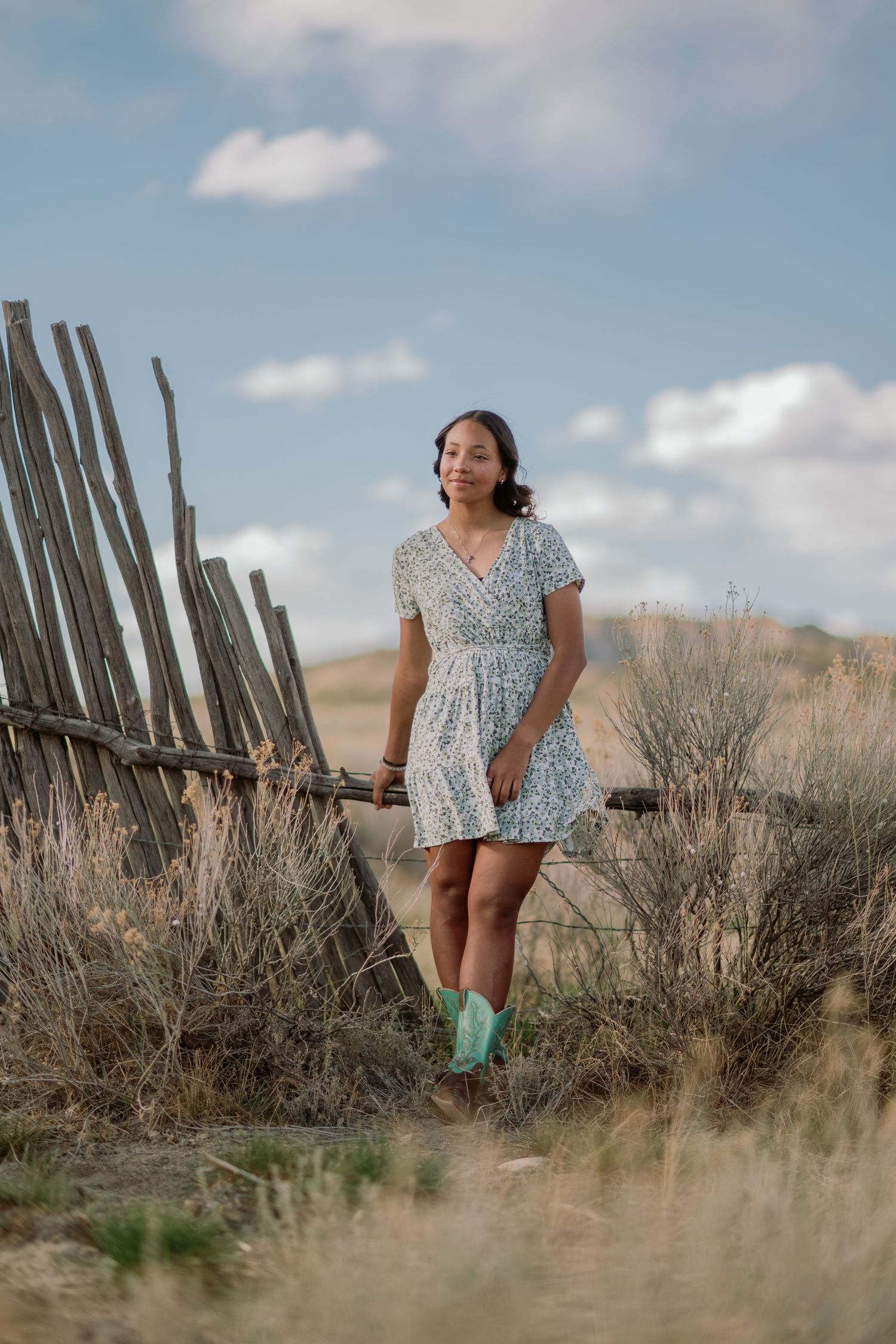 A person in a floral dress and mint green boots stands beside a wooden fence in a rural landscape with cloudy skies.