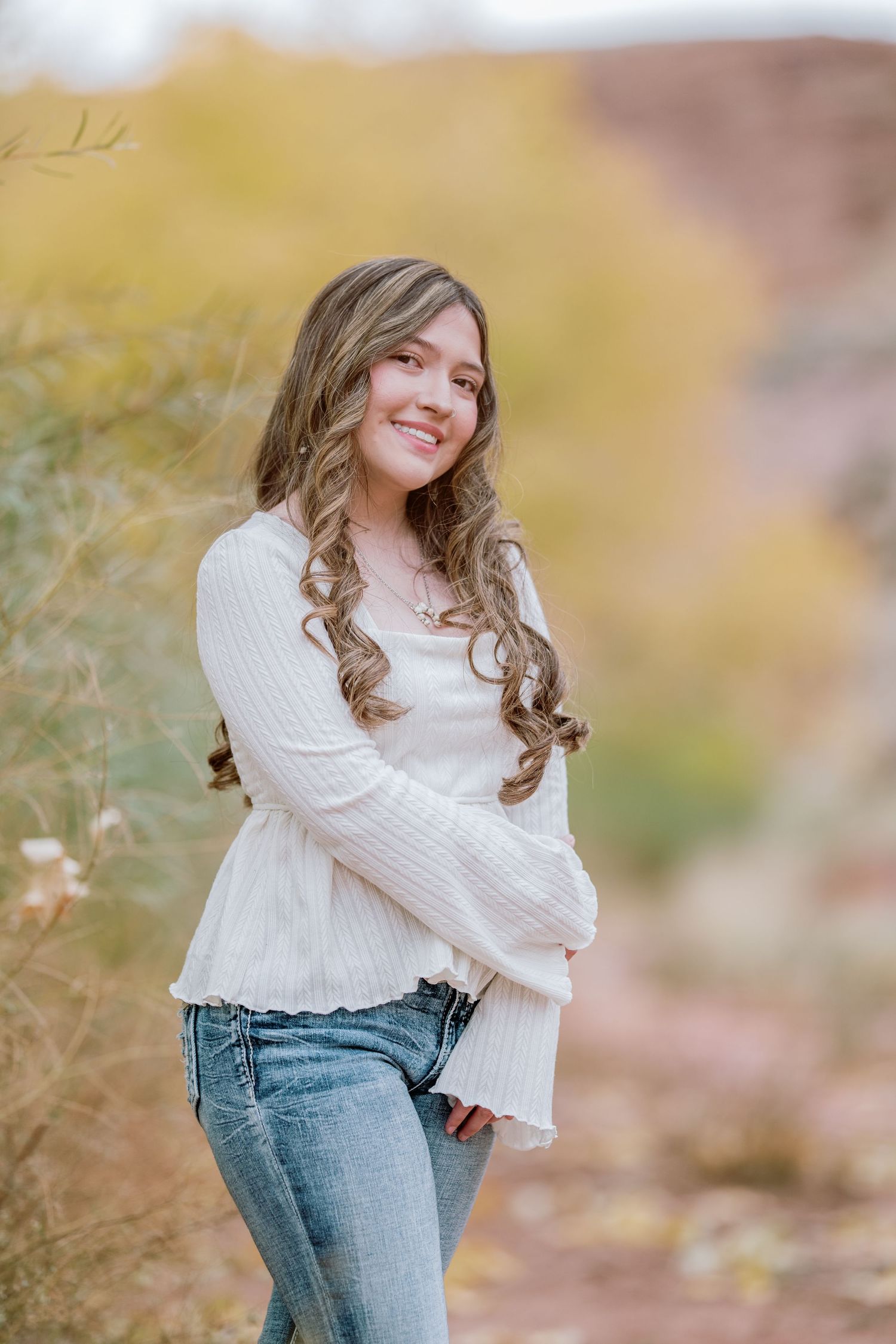 A person wearing a cozy white sweater and blue jeans poses outdoors against a soft autumn background.