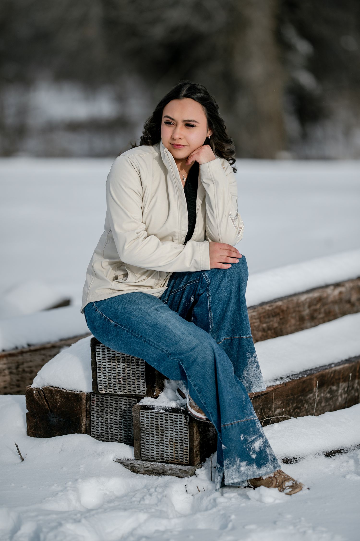Person in cream sweater and blue jeans sits thoughtfully on wicker basket in snowy winter landscape.