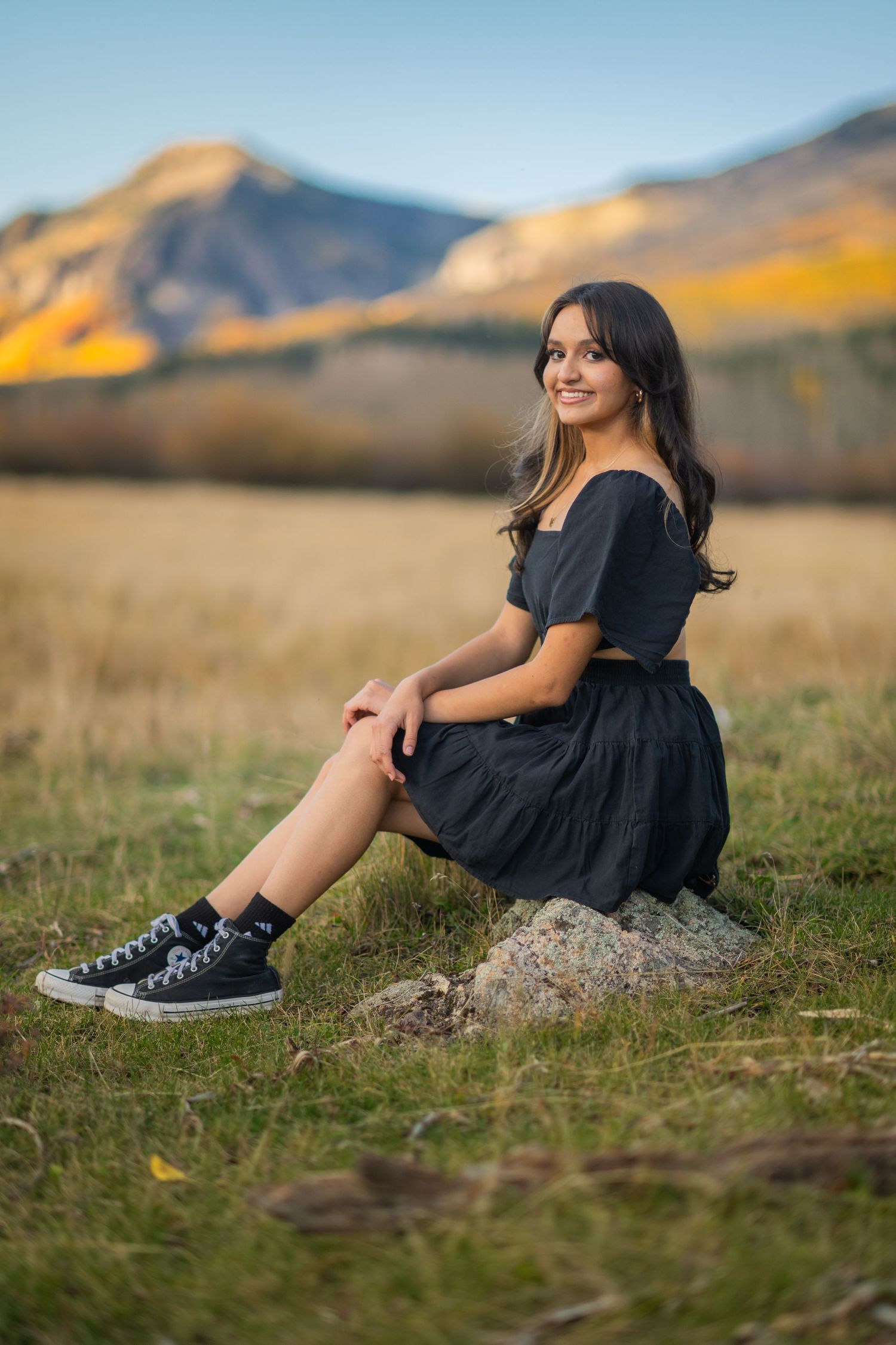 A person in a black dress and sneakers sits on a rock in a scenic mountain meadow during golden hour.