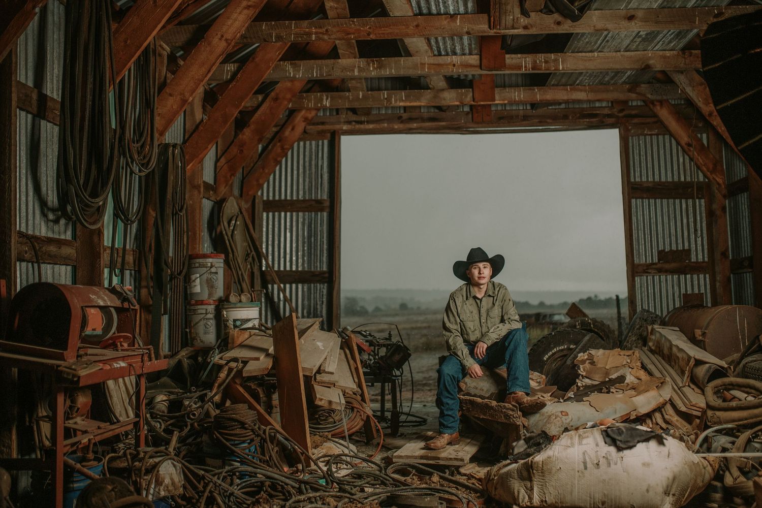 A rustic barn interior scene with western wear and weathered farm equipment amidst hay bales and wooden beams.