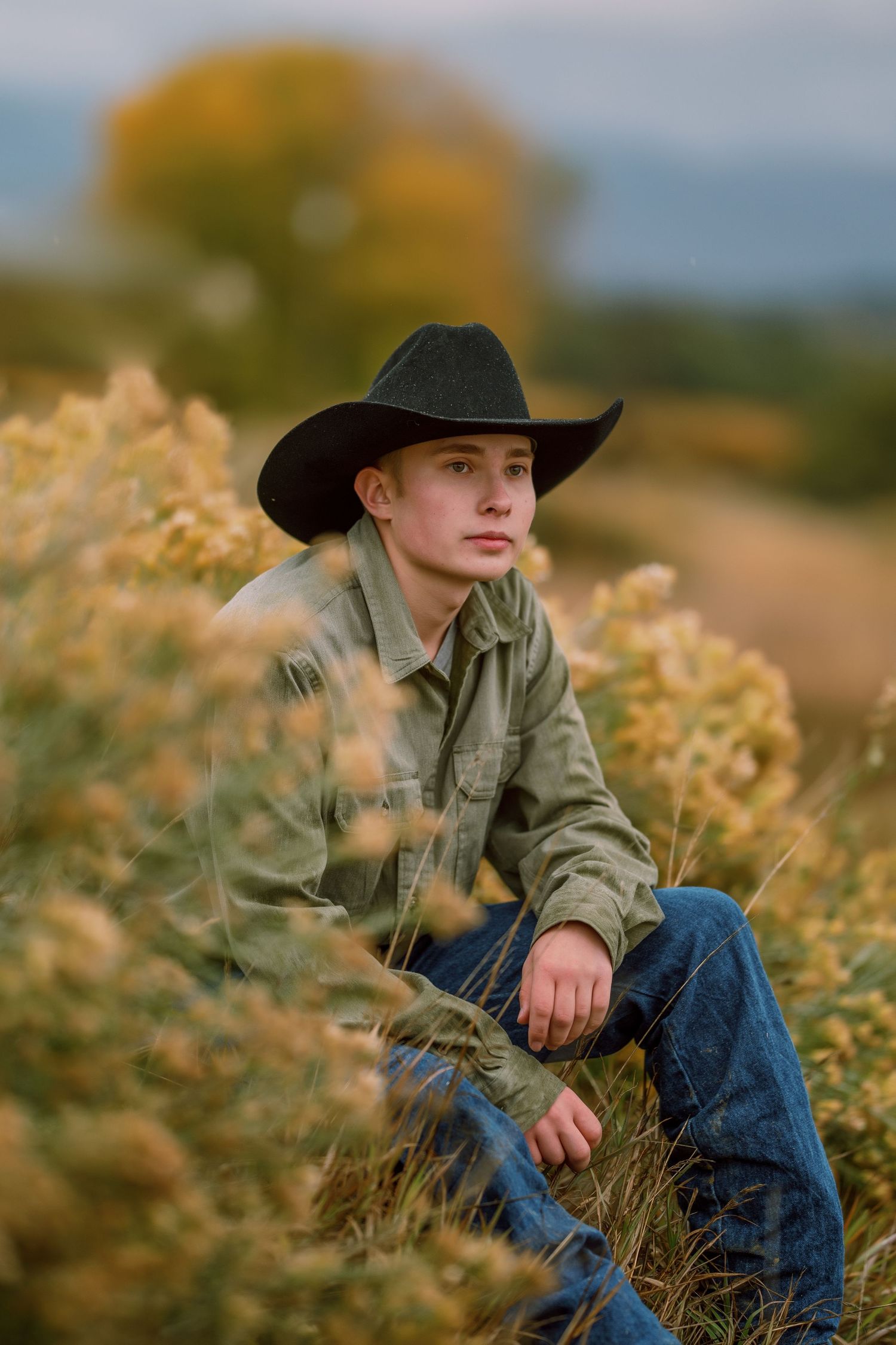 A figure in a black hat and olive jacket sits thoughtfully among golden autumn wildflowers in a rural field at sunset.