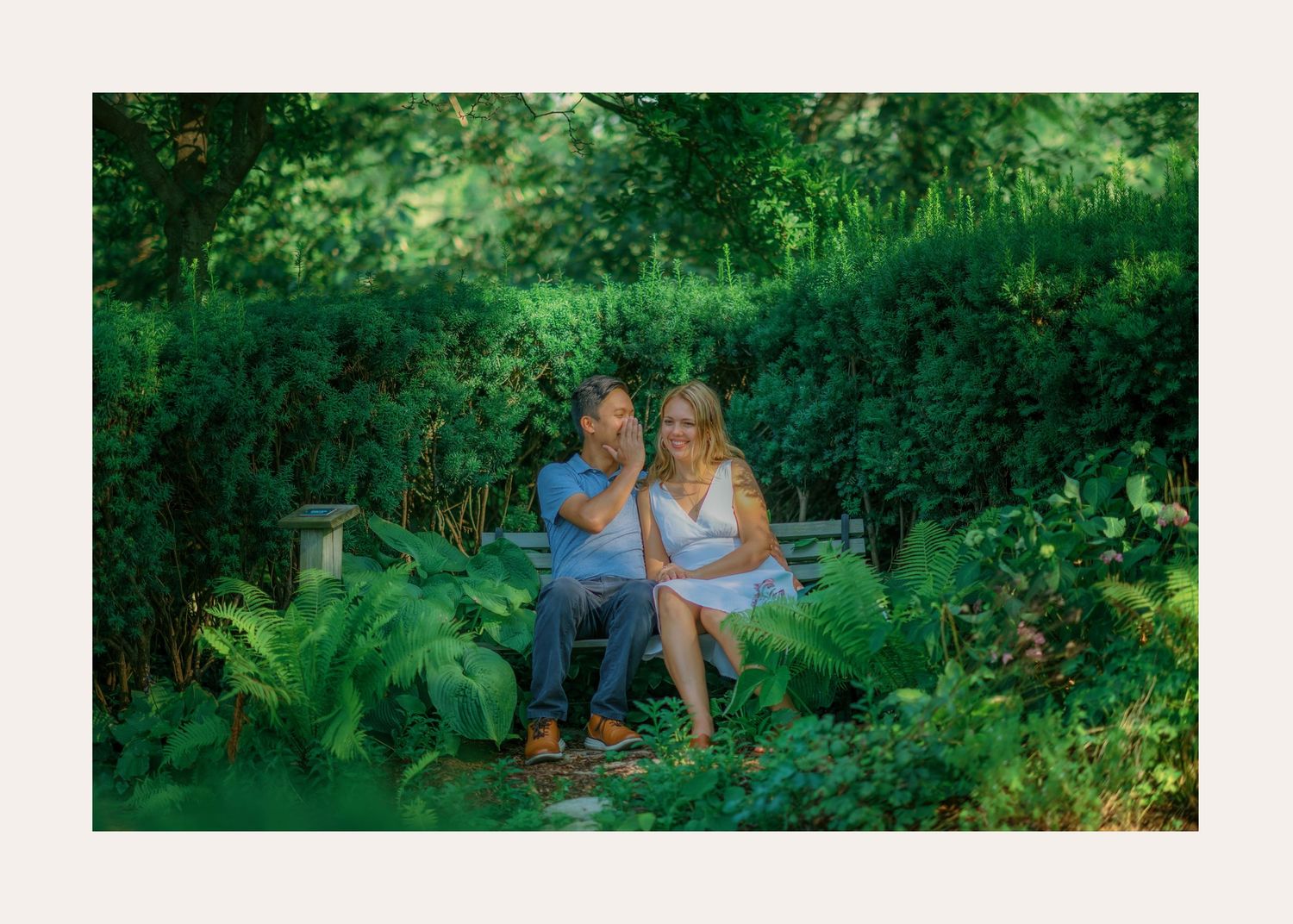 Two people sit together surrounded by lush greenery and flowers in a garden setting.