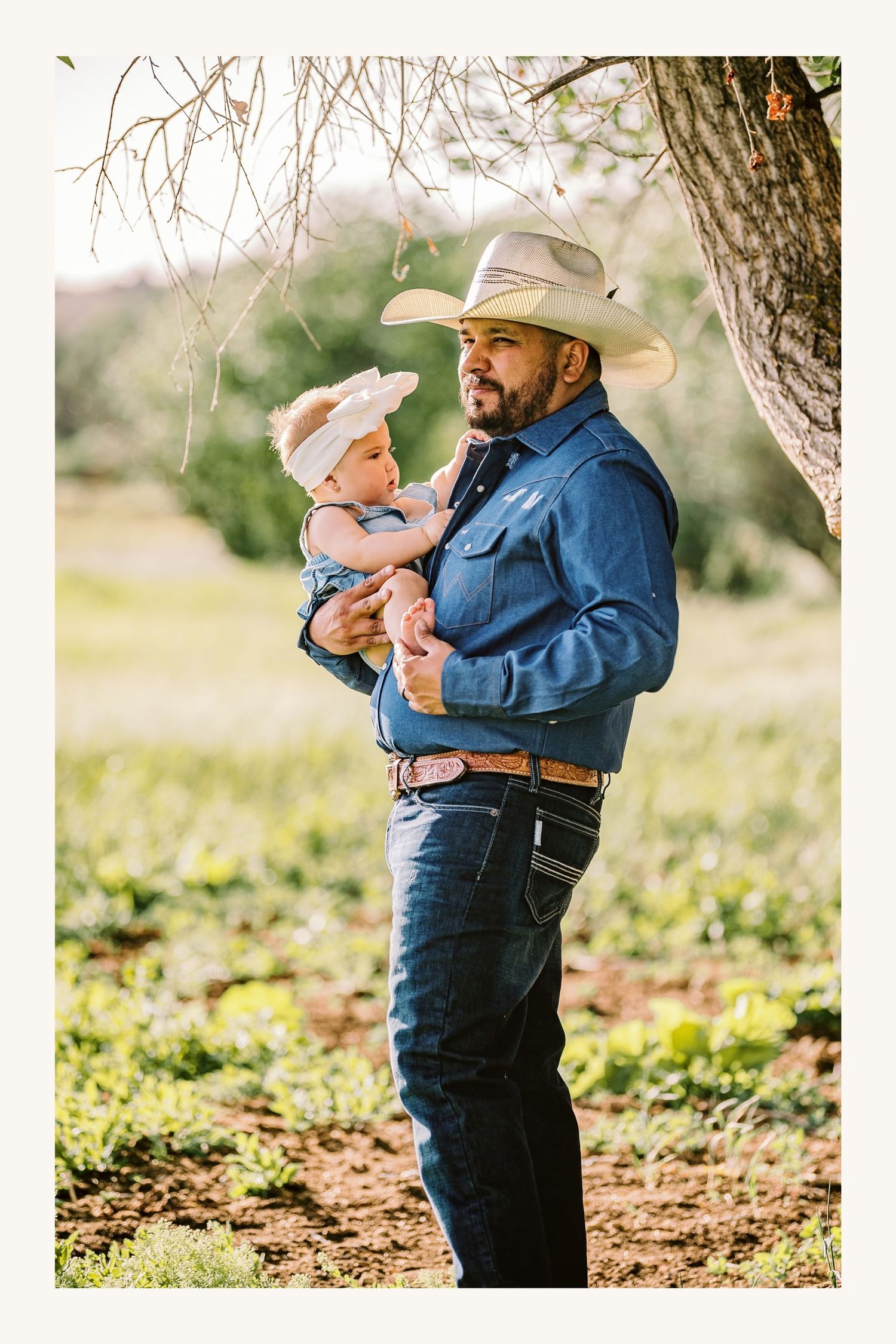 A cowboy father in a blue shirt and white hat lovingly holds his baby outdoors near a tree.