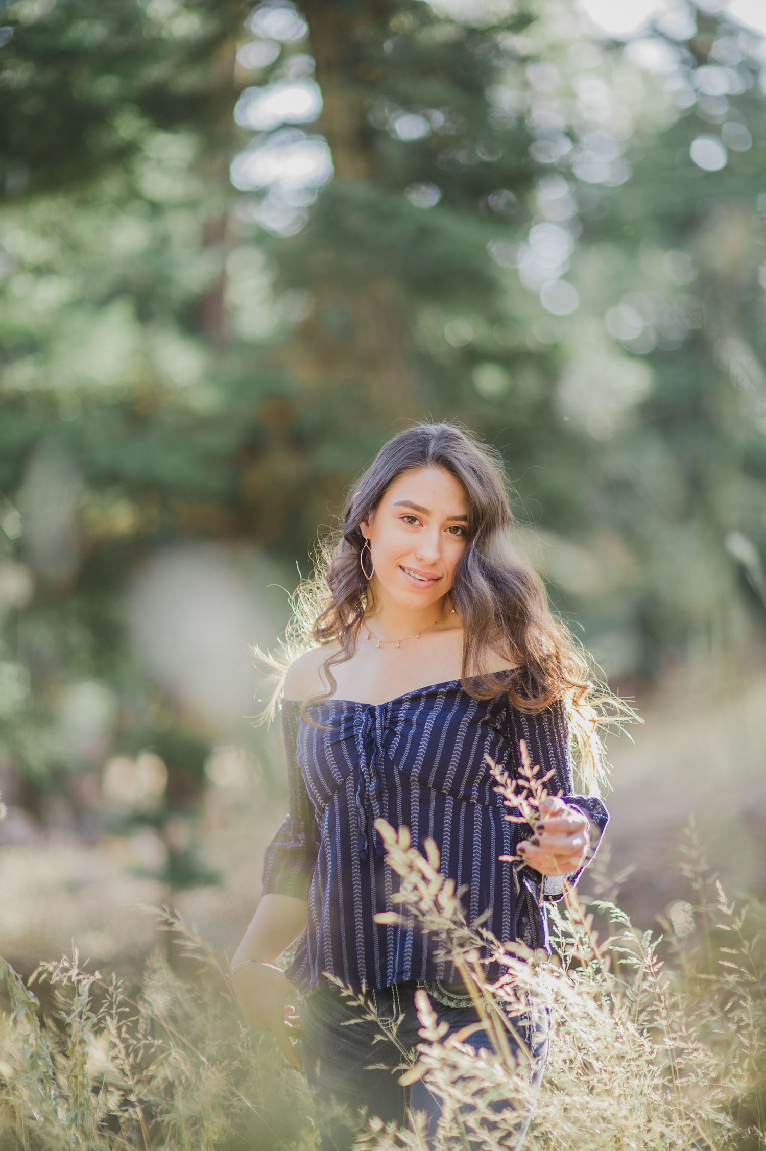 Young woman in off-shoulder striped top standing in sunlit forest meadow holding wildflowers.