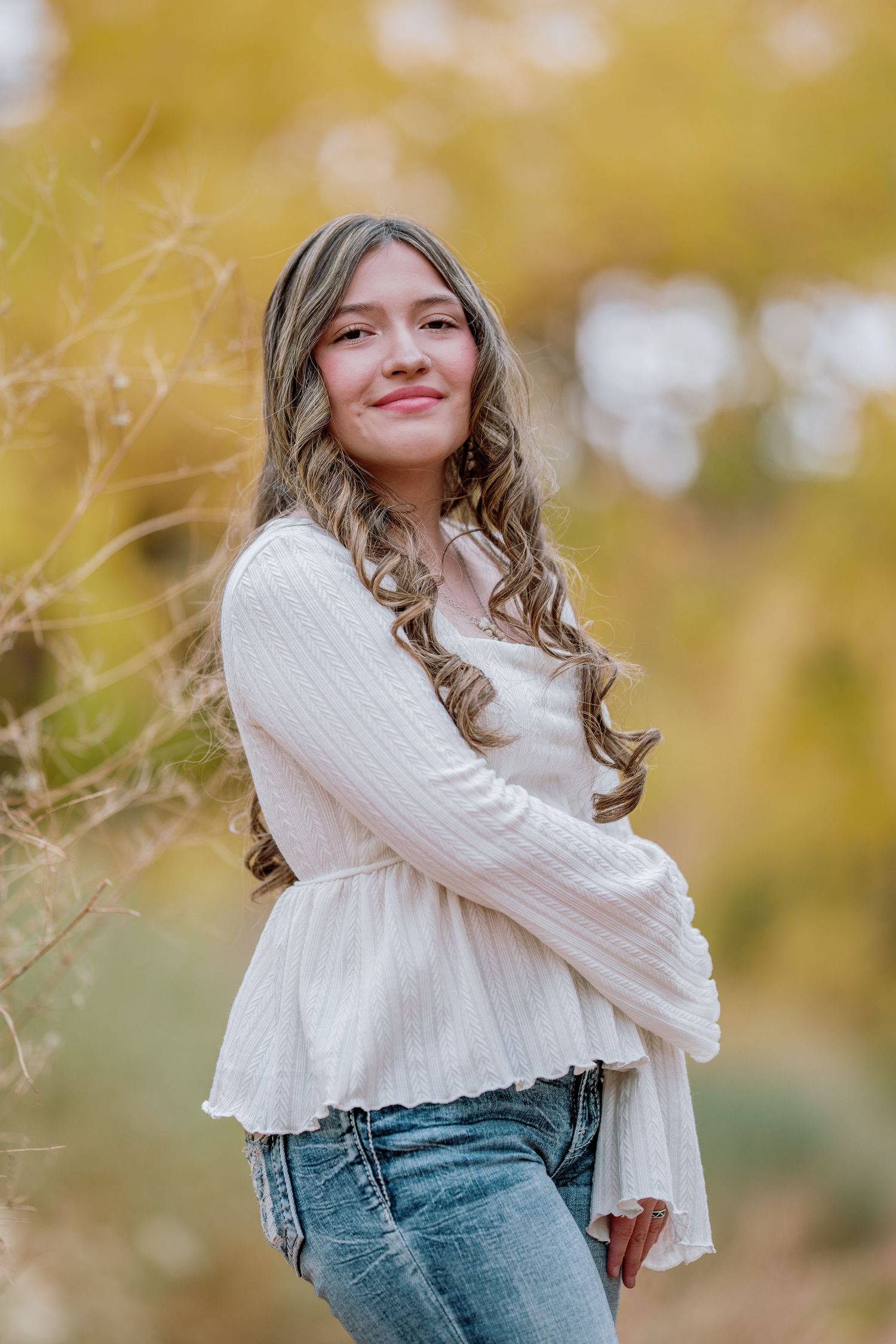 A person in a white sweater and jeans poses outdoors against a soft autumn background with blurred golden bokeh.