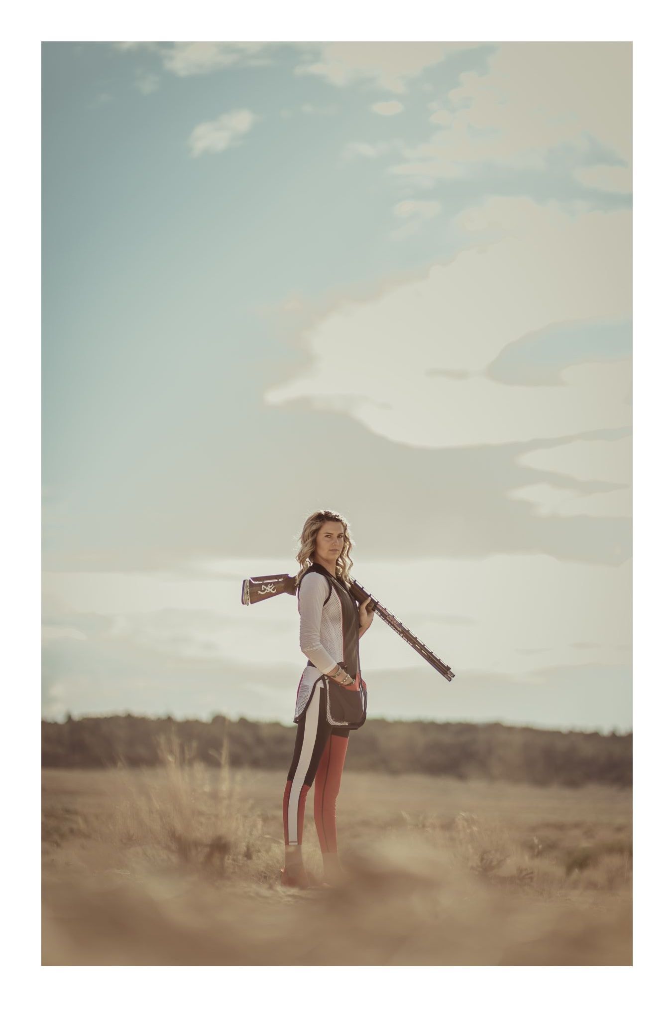 A young woman stands in an open field holding a rifle over her shoulder, wearing a white jacket and red pants.