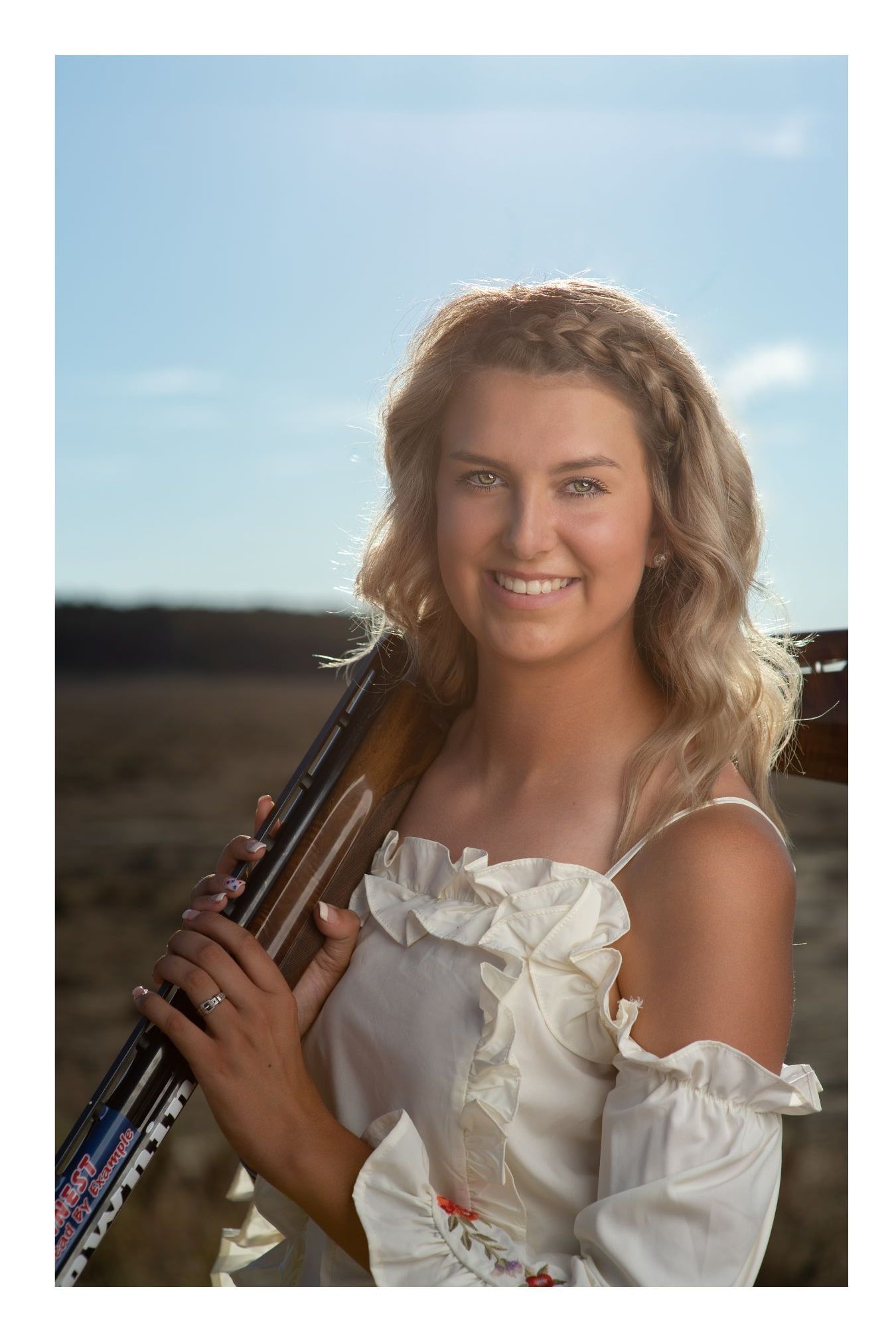 Young blonde woman in white off-shoulder dress holding a rifle, smiling outdoors under a blue sky.
