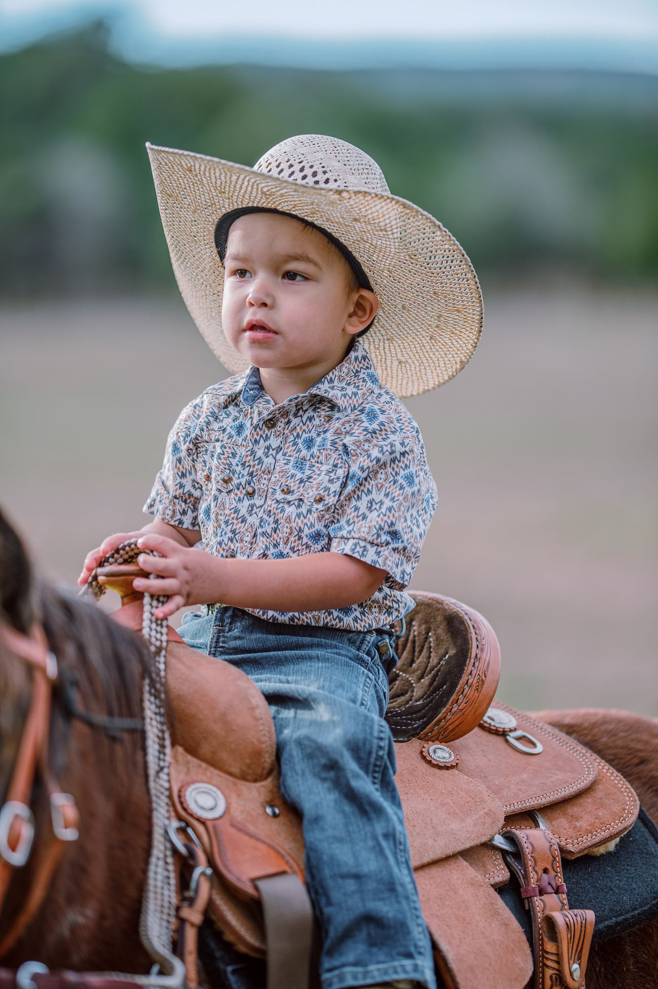 A young boy in a straw cowboy hat and floral shirt sits atop a brown horse with a western saddle outdoors.