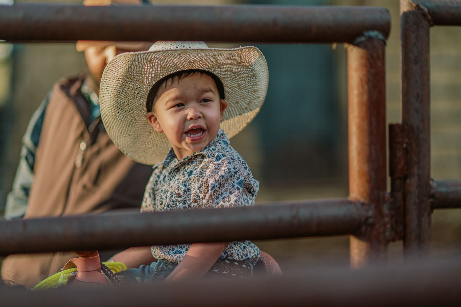 Smiling toddler wearing a straw cowboy hat sitting on a fence rail at a rodeo event.