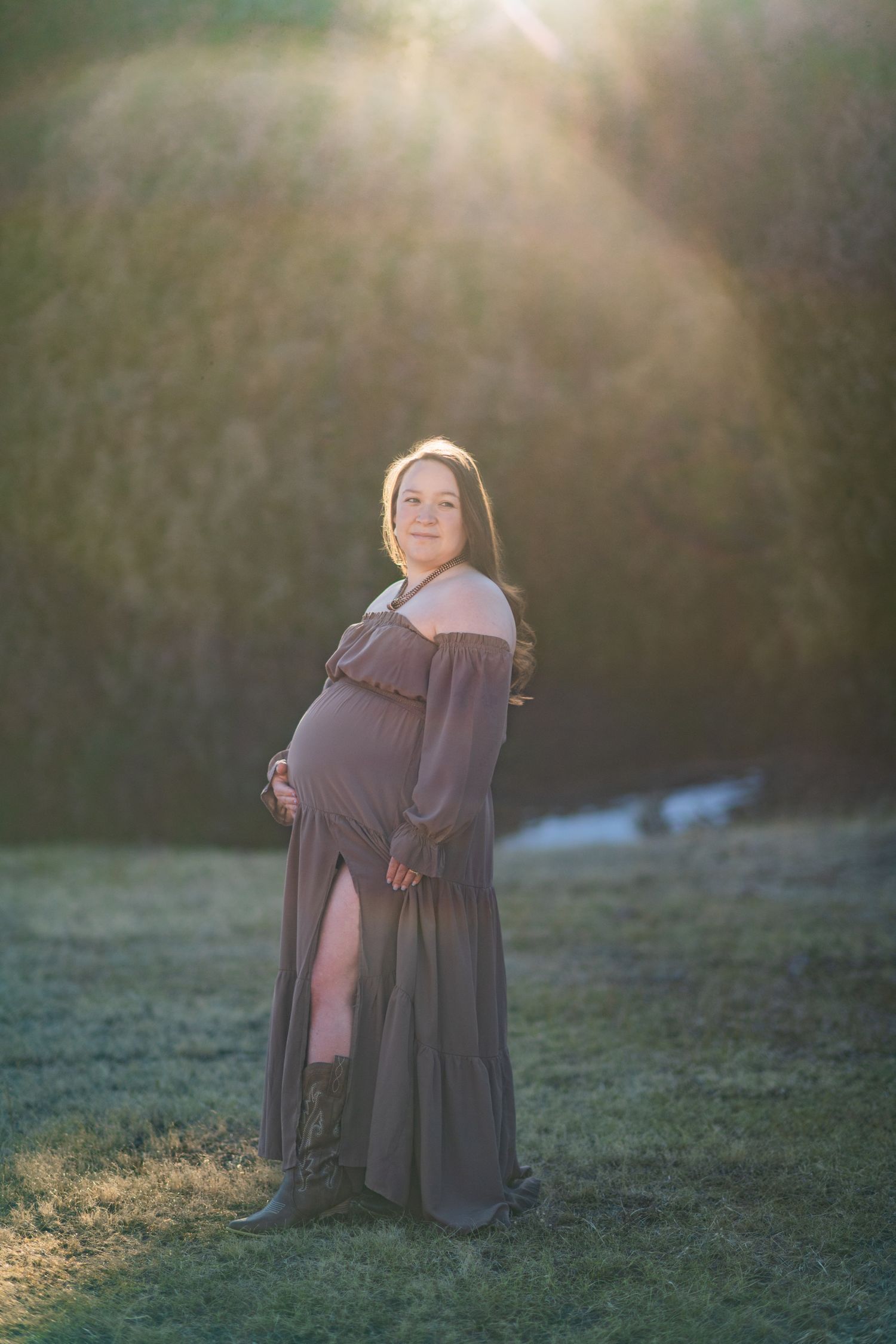 A pregnant woman in a flowing brown dress poses outdoors in soft golden sunlight during a maternity photoshoot.