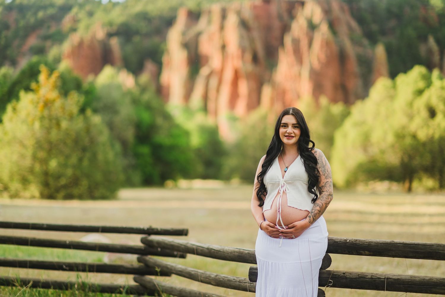 Smiling pregnant woman in white dress poses outdoors near rustic fence with red rock formations and green trees behind her.