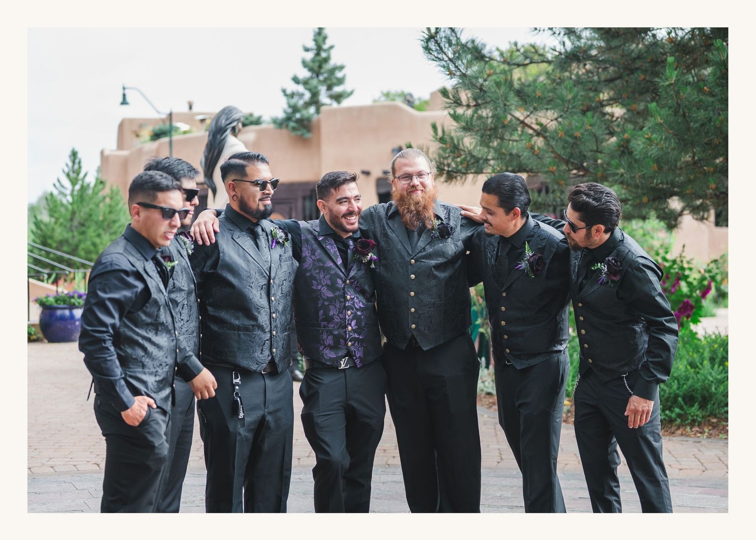 Six men in black suits smiling and posing together outdoors at a wedding venue surrounded by trees.