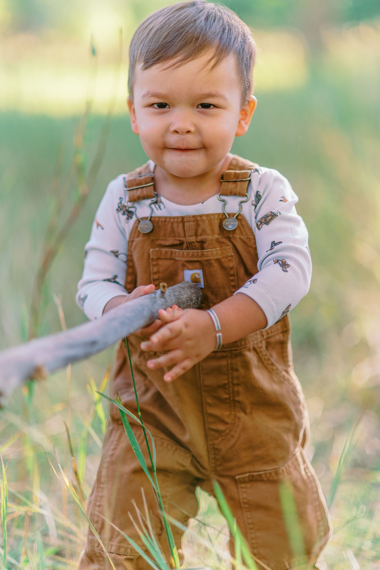 Small child in brown overalls plays outdoors in a grassy field on a sunny day.