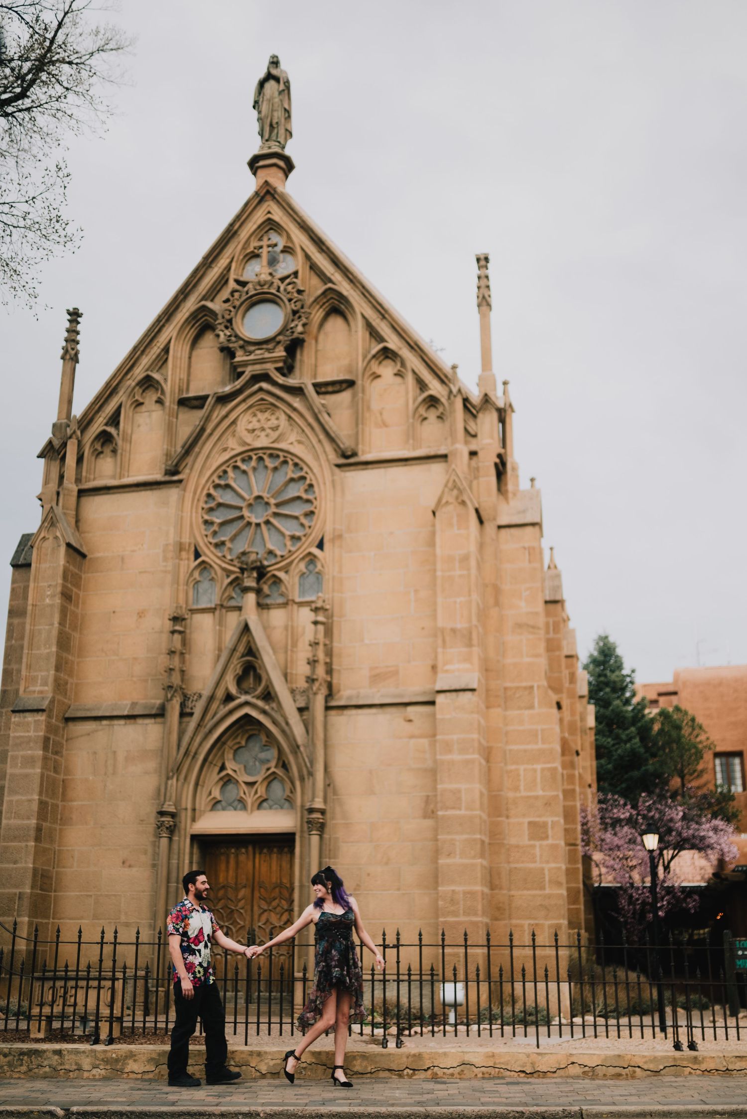 Couple holding hands in front of historic Gothic stone chapel with rose window and statue on overcast day.