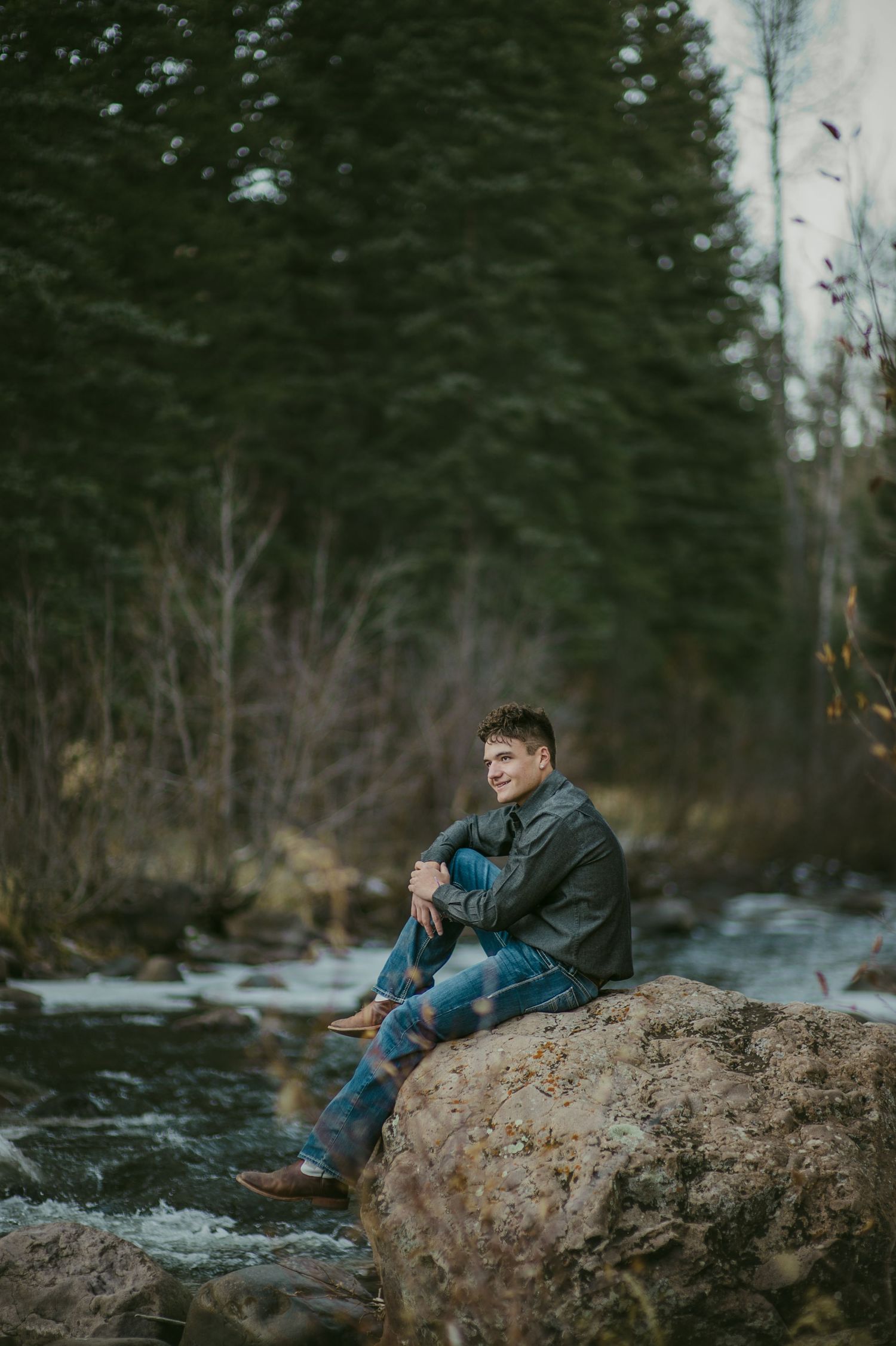 Young man sitting on a rock by a mountain stream, wearing a leather jacket and jeans, surrounded by evergreen trees.