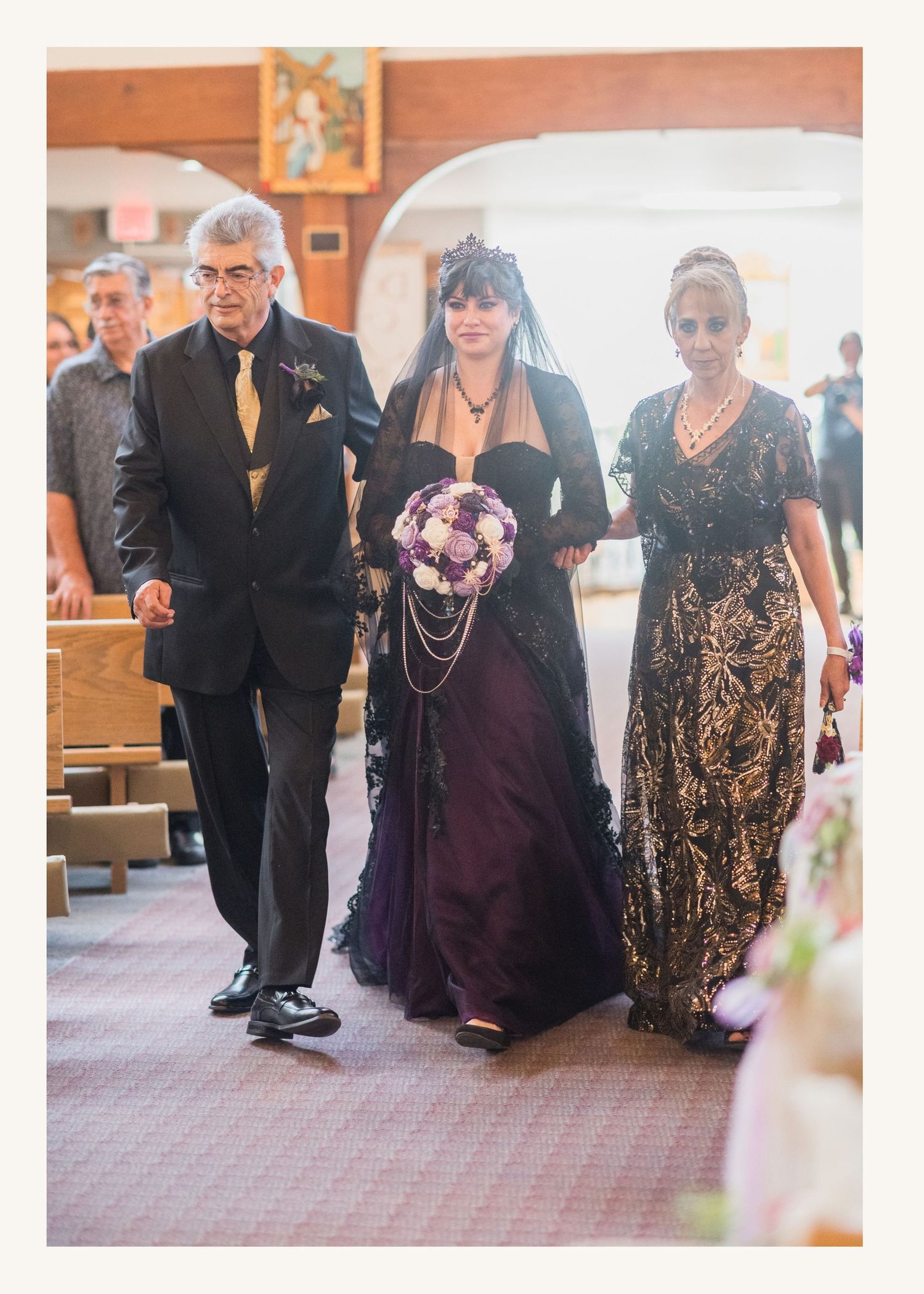Bride in black and purple gown walks down church aisle with parents during wedding ceremony.