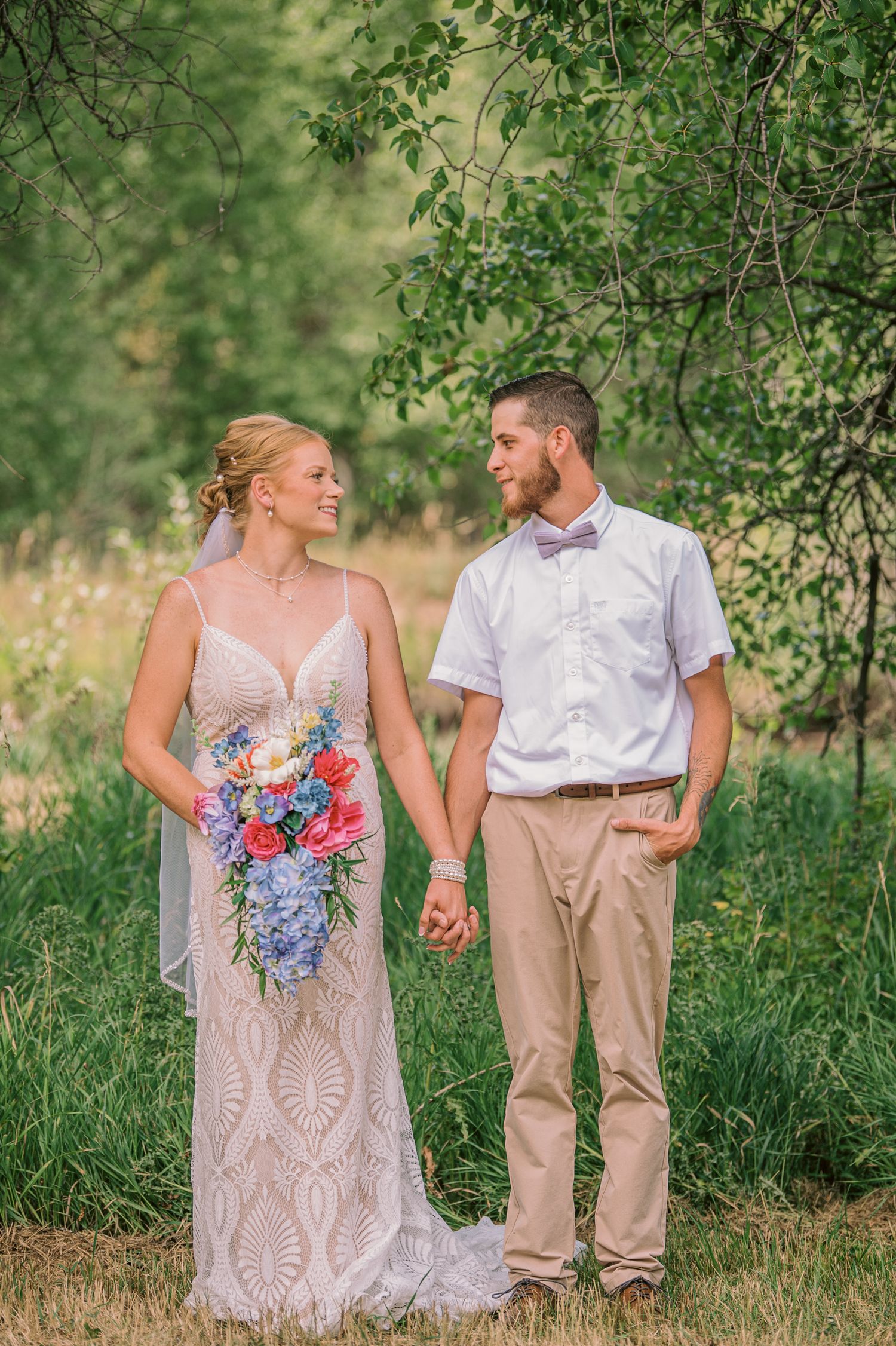 Bride and groom holding hands in a lush green outdoor setting, bride holding colorful bouquet in elegant lace gown.