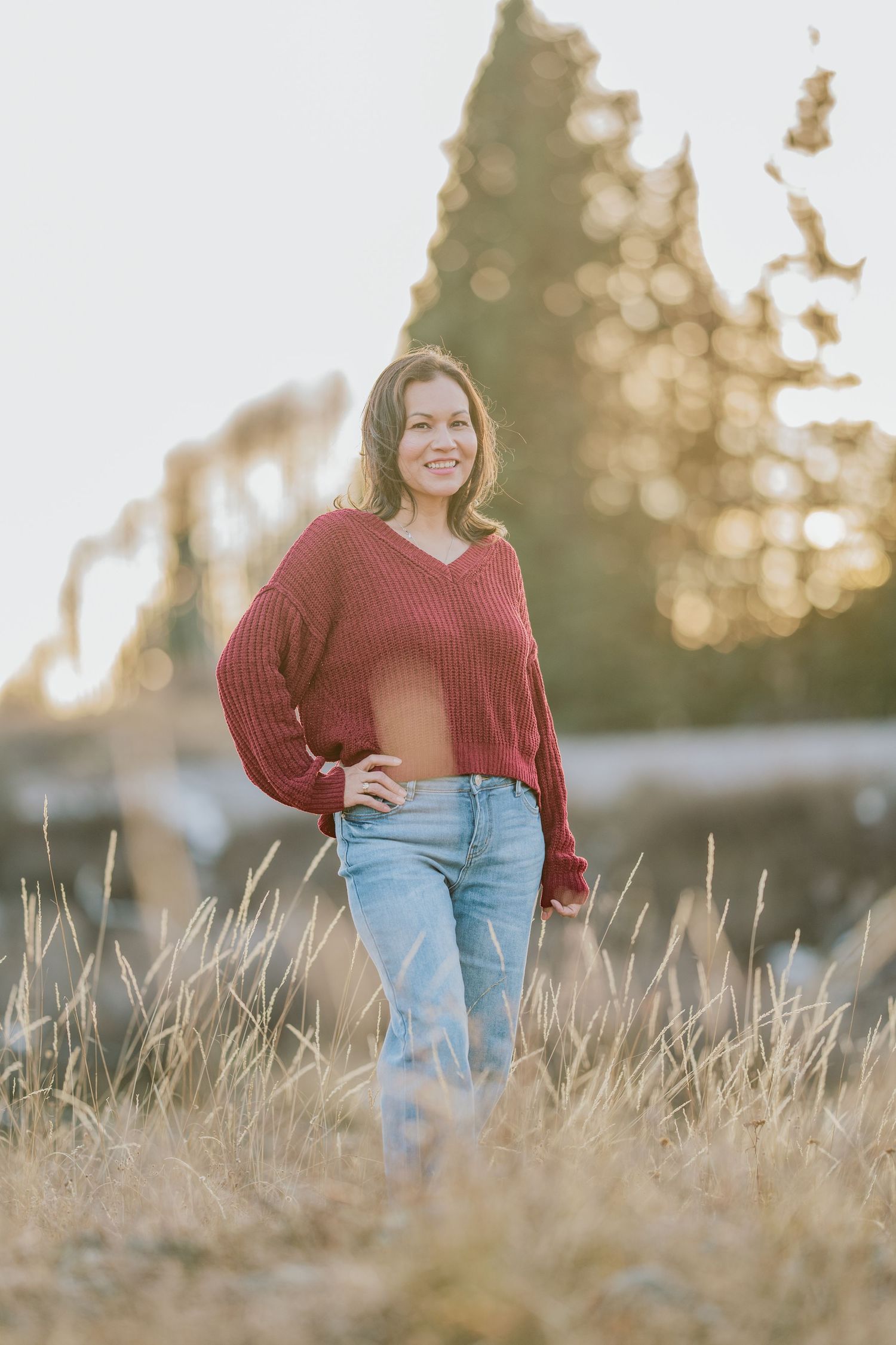 A person in a red sweater and jeans stands in a field of tall grass at golden hour with bokeh lighting effects.