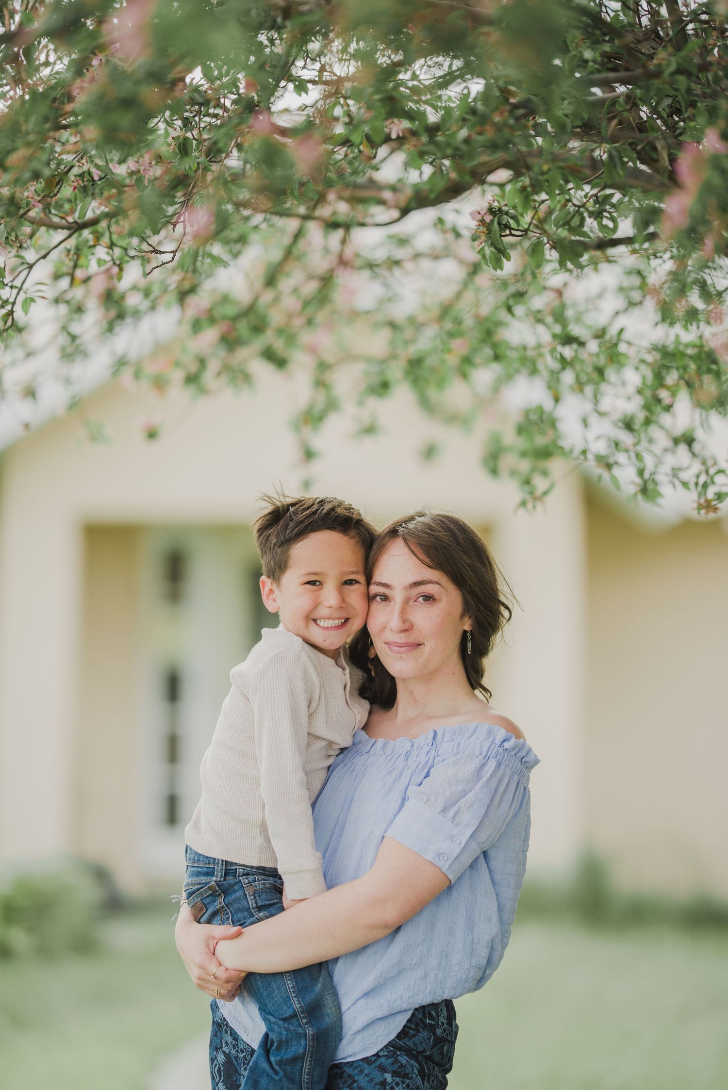 A parent and child embrace under blossoming tree branches in a springtime outdoor portrait.