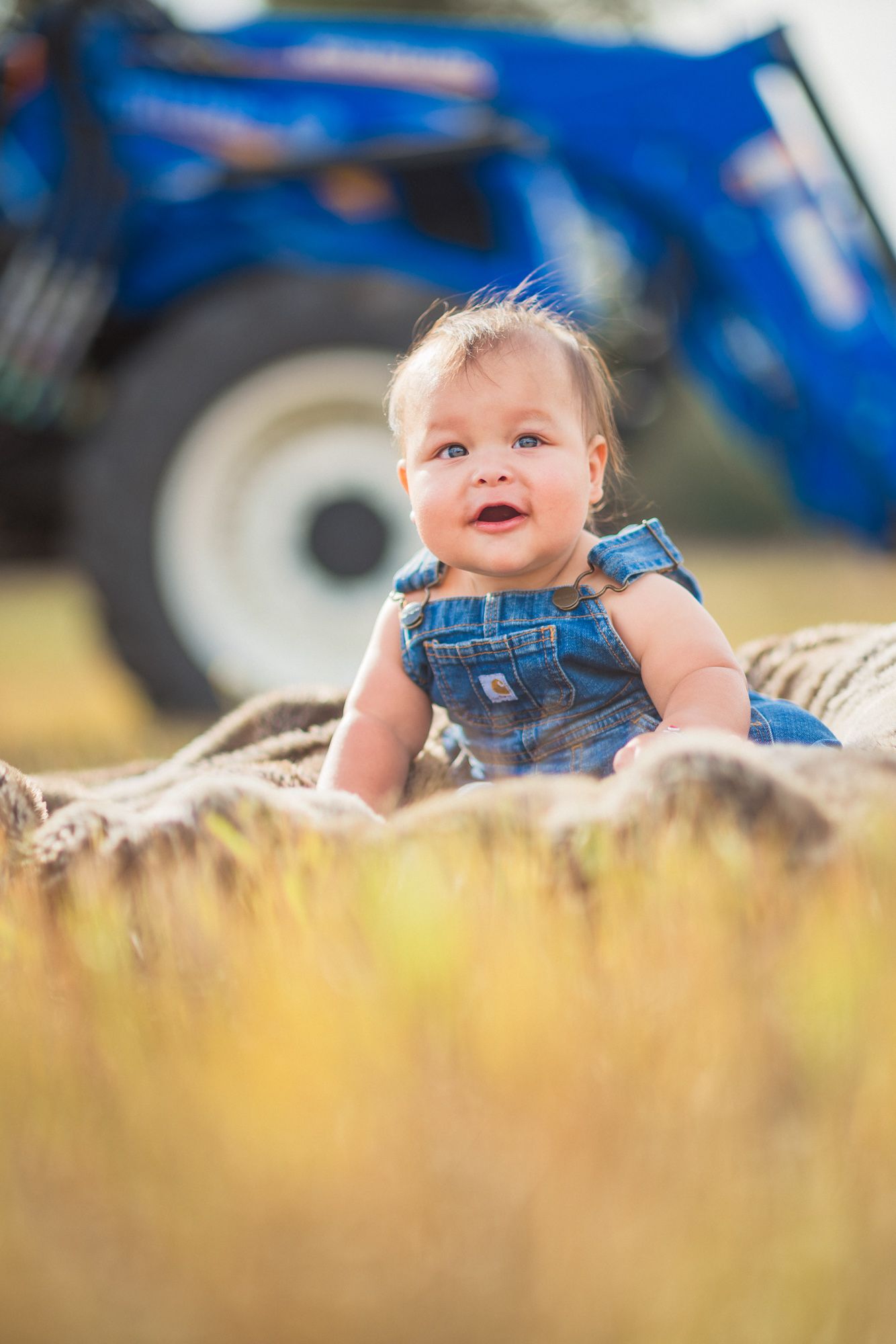 Smiling baby in denim overalls sitting on a hay bale in a sunny field with a blue tractor in the background.