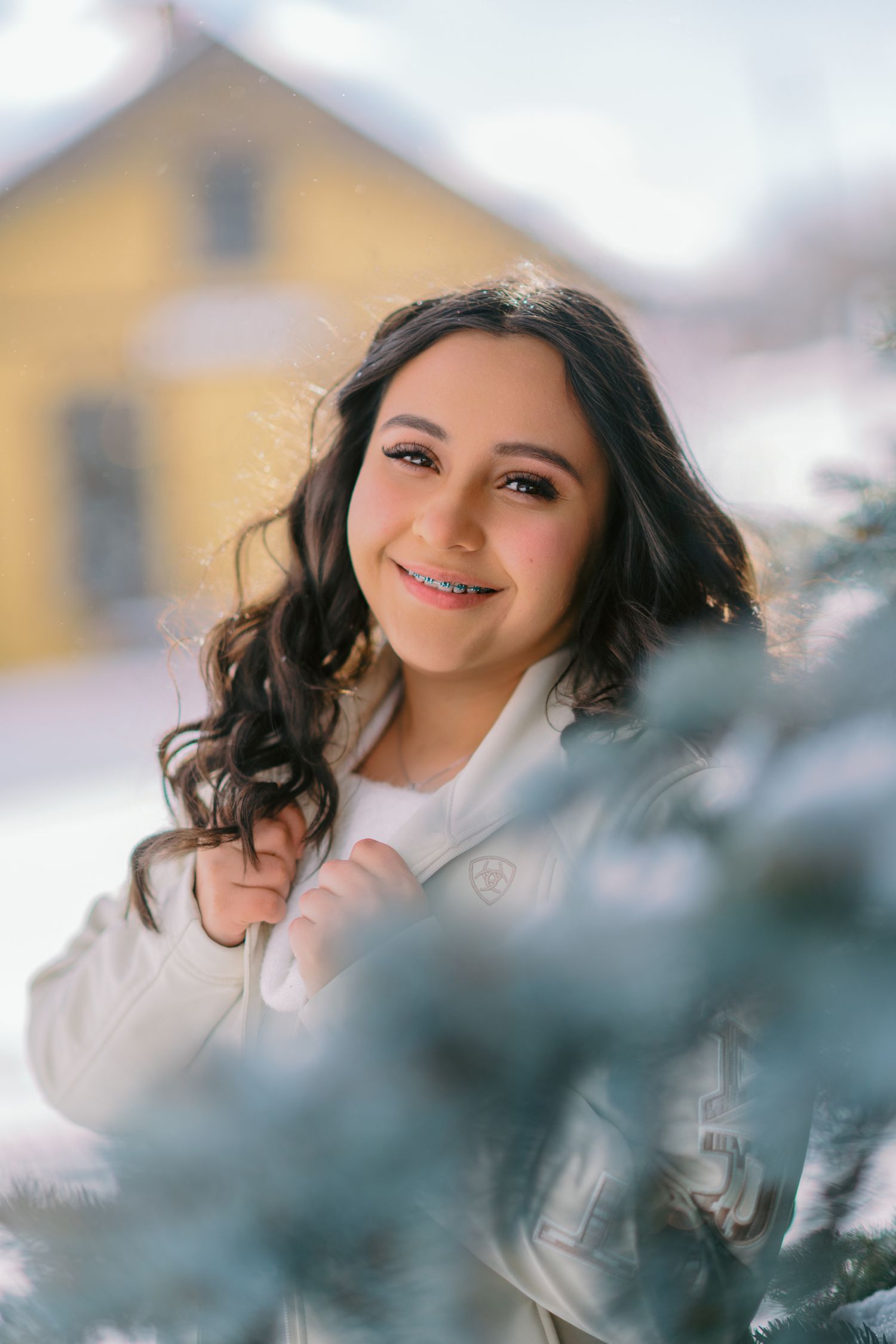 A person wearing a white sweater poses outdoors with evergreen branches in the foreground creating a dreamy winter scene.