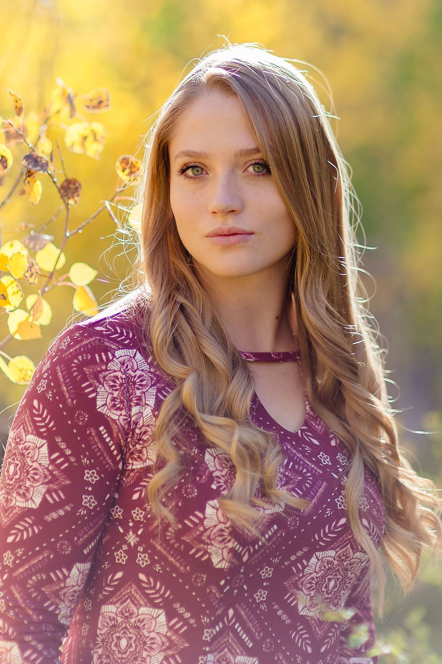 Portrait in warm sunlight wearing a burgundy floral pattern top with long wavy blonde hair against yellow autumn foliage.